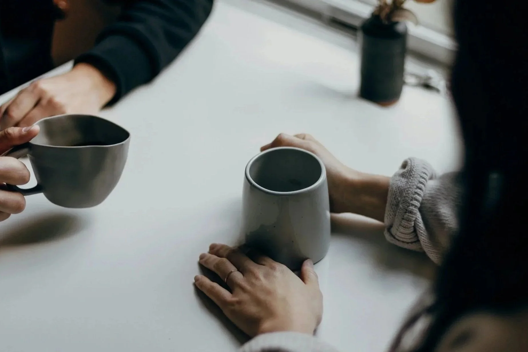 Two people holding ceramic coffee mugs at a white table in a cozy indoor setting.