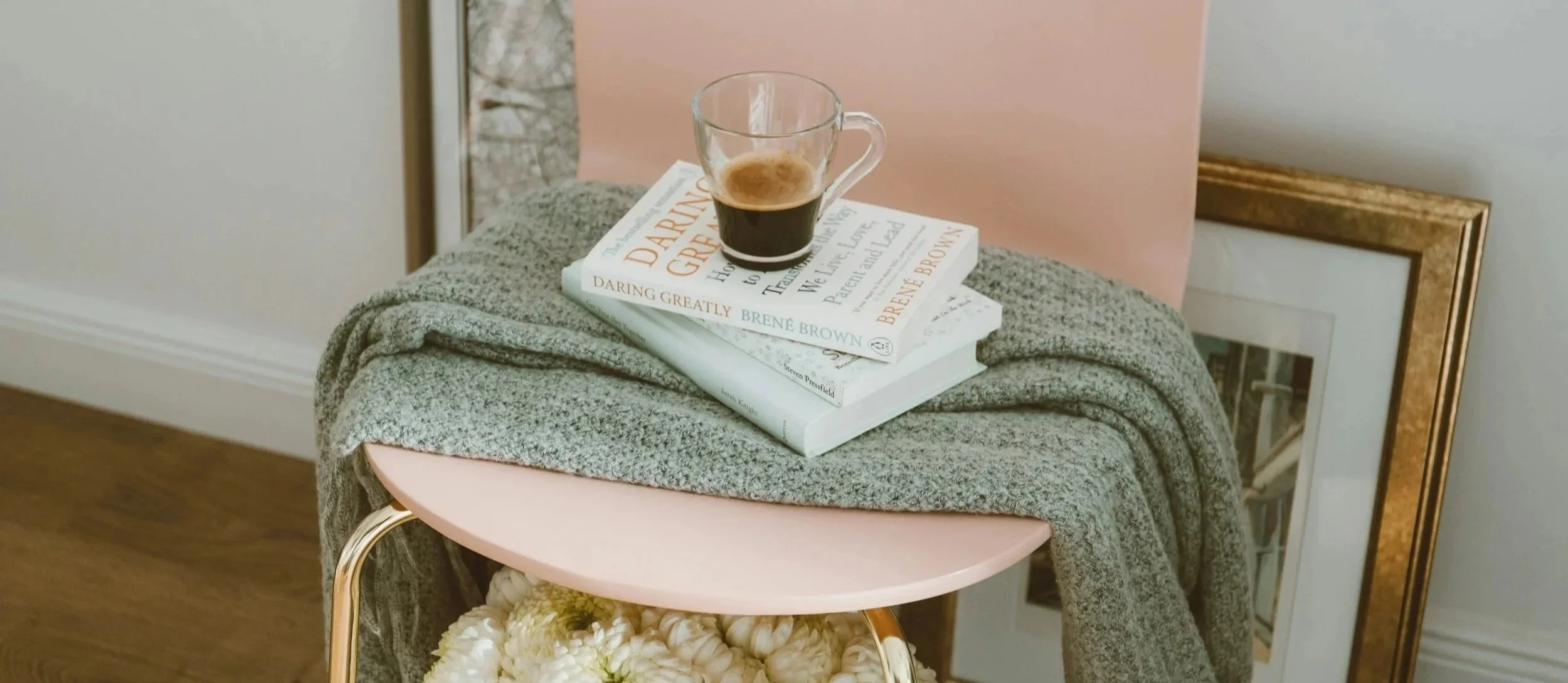 A pink chair with a gray blanket draped over it, a stack of three books, and a glass of coffee on top. The books are titled "Daring Greatly" by Brené Brown, "The Gift of Imperfection" by Brené Brown, and "The Light After the War" by Anita Abriel. The chair is next to a framed picture leaning against the wall.