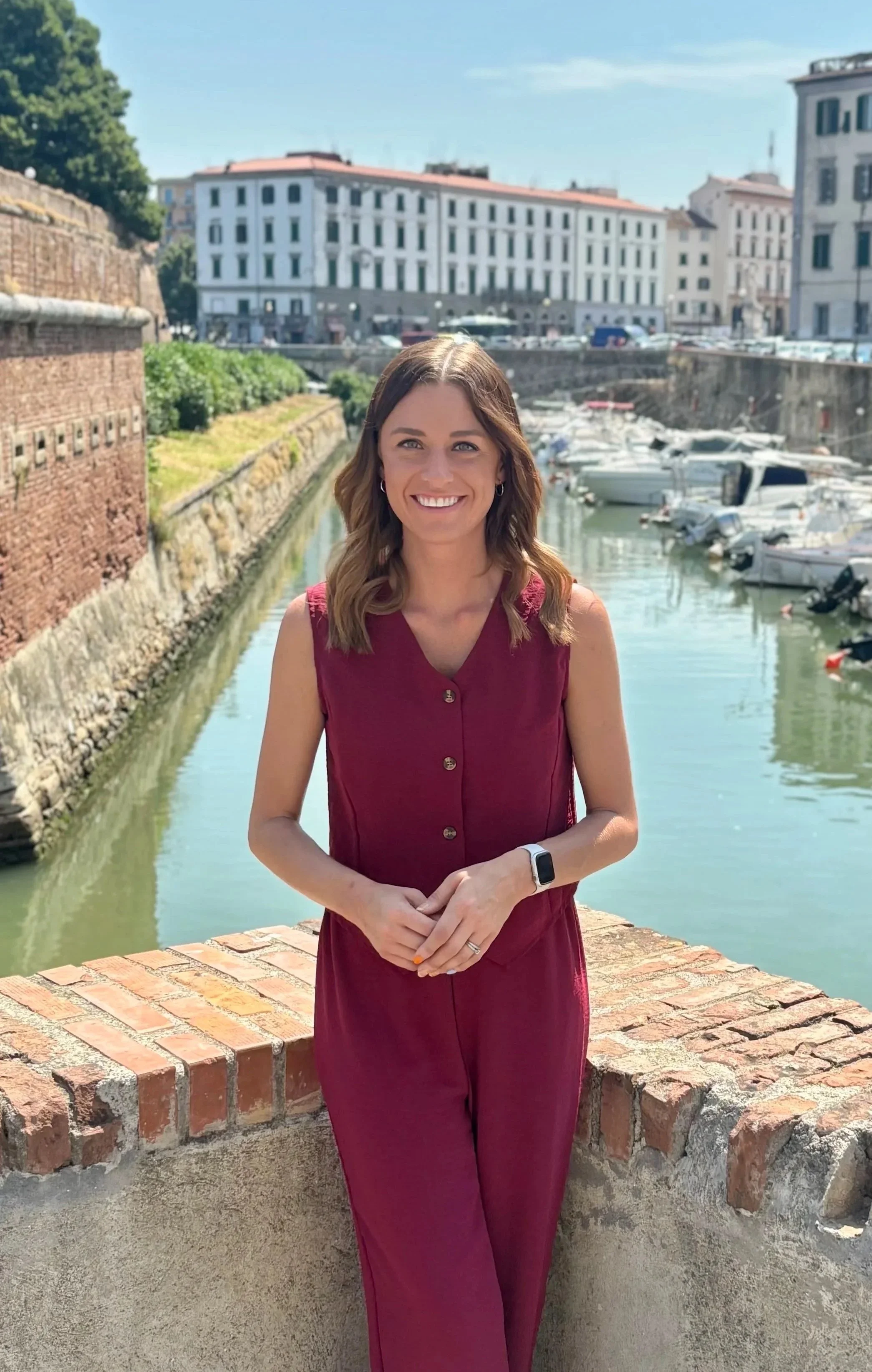A woman with wavy brown hair smiling and wearing a sleeveless burgundy outfit, standing on a brick ledge by a canal with boats and historic buildings in the background, on a sunny day.