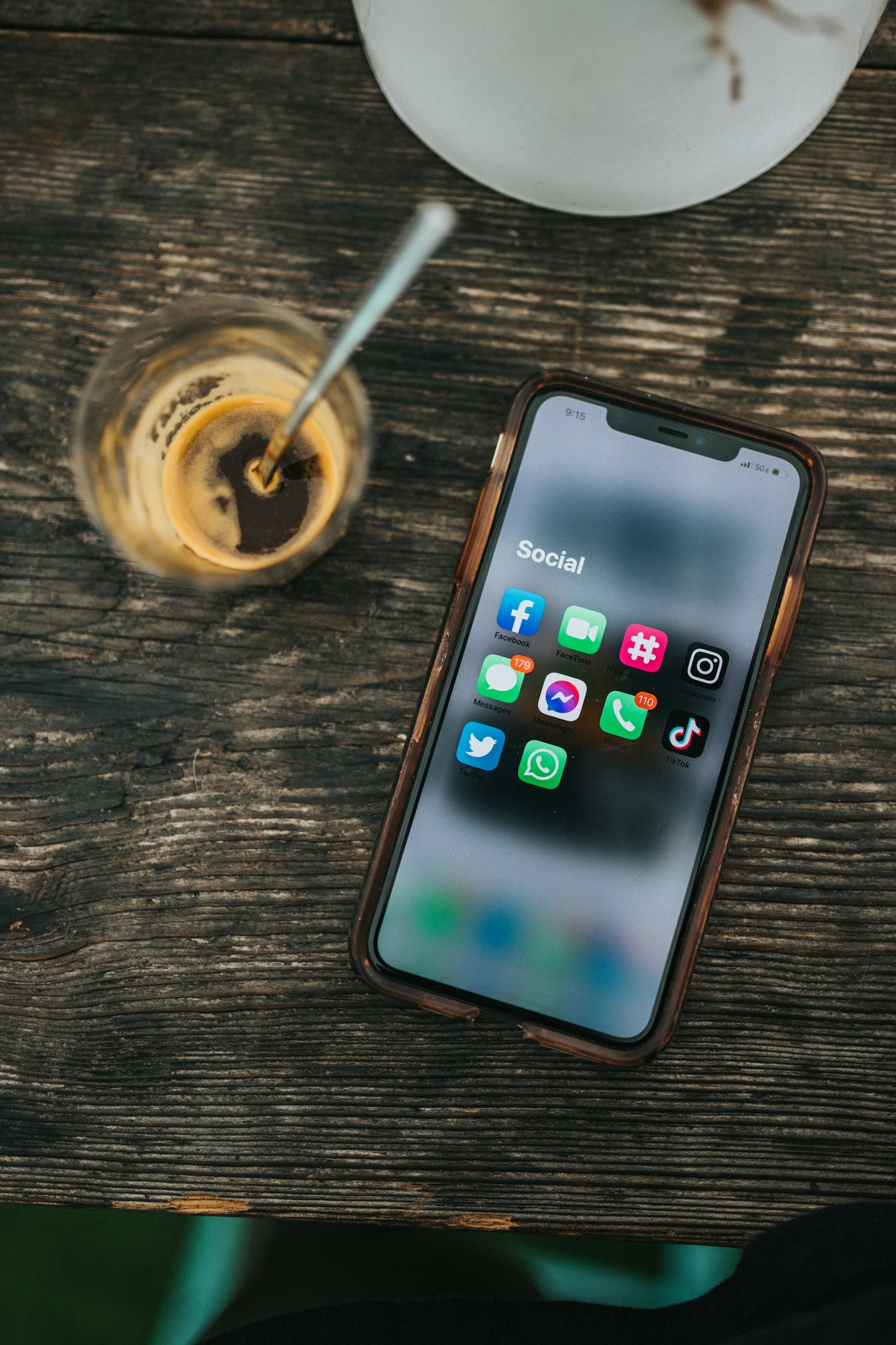 Smartphone on a wooden table displaying social media apps, a partially consumed espresso in a glass with a straw, and part of a white bowl.