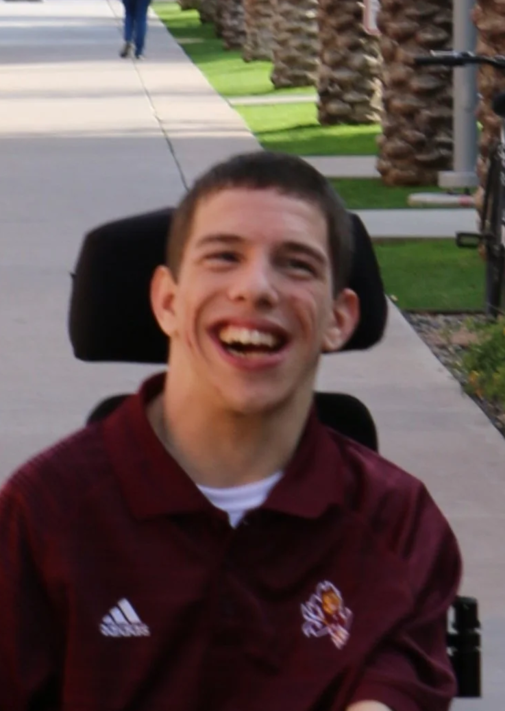 A young man with short dark hair smiling outdoors, sitting in a wheelchair, wearing a maroon sports jacket with a logo.