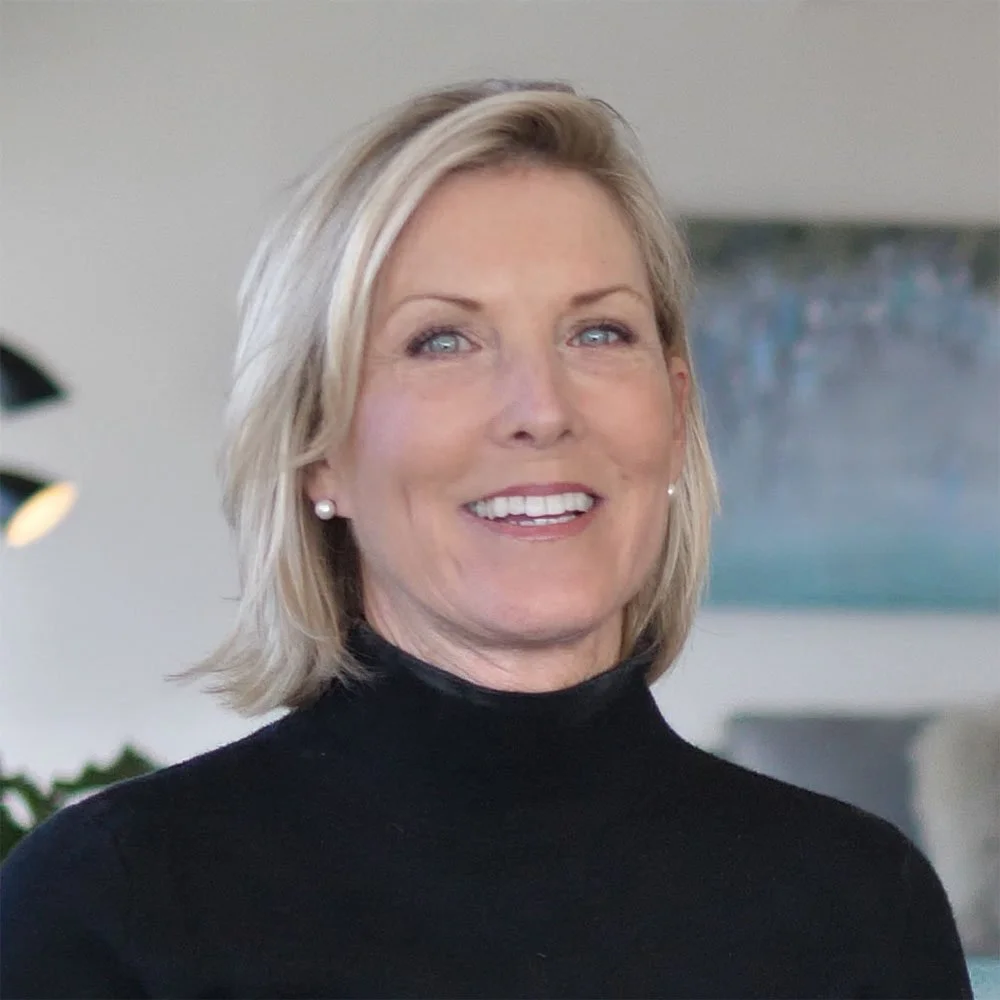 Close-up image of a middle-aged woman with blonde hair, wearing pearl earrings and a black turtleneck sweater, smiling in an indoor setting.