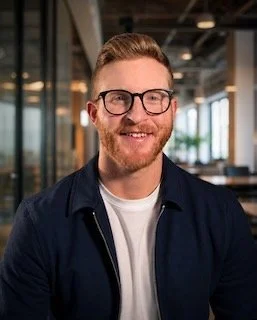 Portrait of a young man with red hair, glasses, and a beard, wearing a black collared shirt, against a dark background.