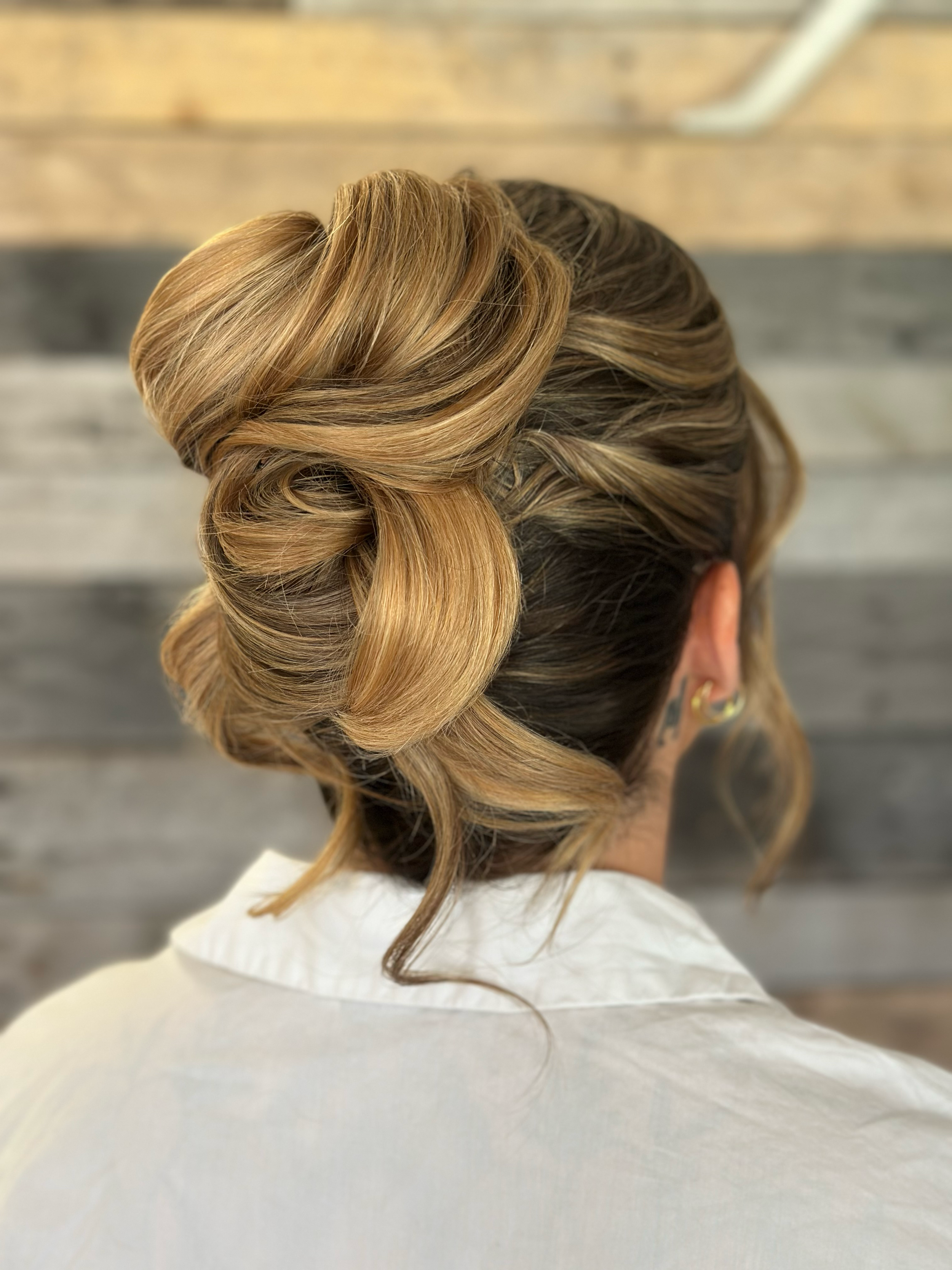 Back of a woman's head with an elegant updo hairstyle featuring large, intricate rolls and waves, and a loose curl hanging down. She is wearing earrings and a white shirt or smock, with a wooden panel wall in the background.