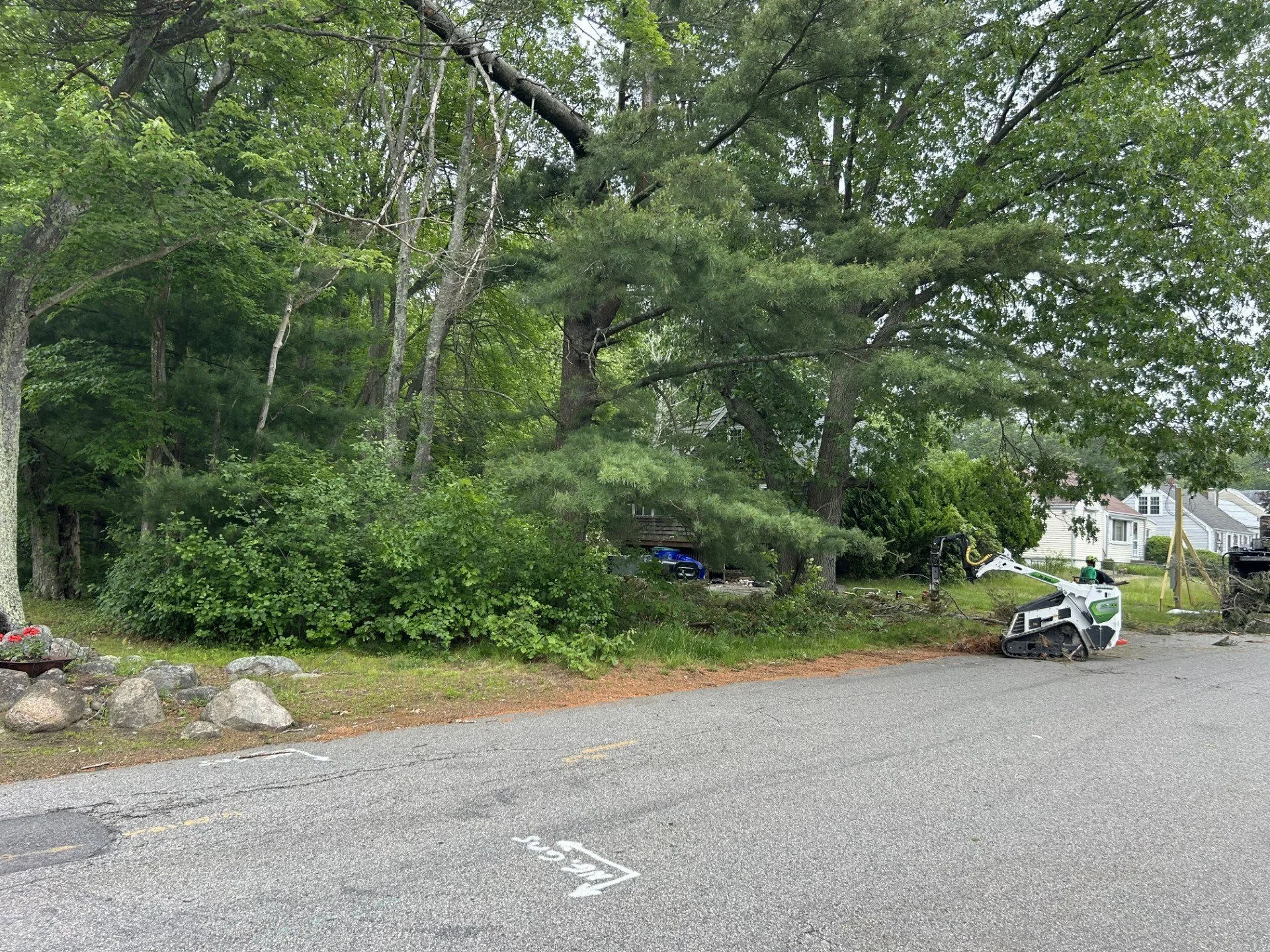 Street with a small track loader digging into the soil near trees on a green yard in a residential neighborhood.