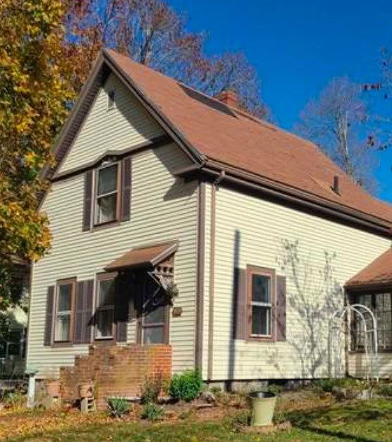 A two-story house with beige siding and brown window shutters, a brick porch, and a pitched roof with reddish shingles, surrounded by trees with fall foliage.