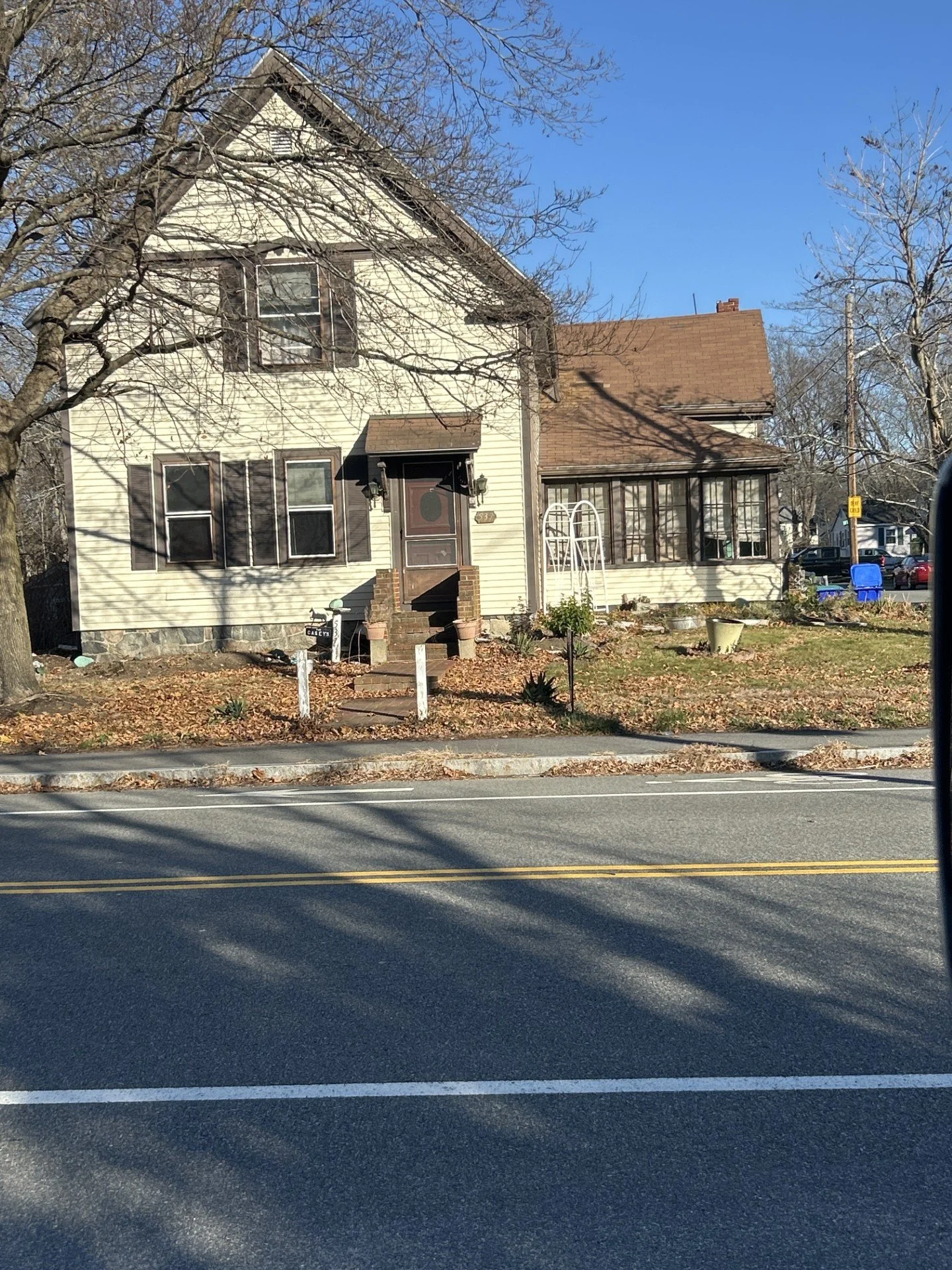 A two-story house with a gabled roof, white siding, and dark window shutters, located on a small yard along a street with double yellow lines. The yard has some plants, mulch, and a small tree with a white arch-shaped support. Bare trees surround the house, and a clear blue sky is visible above.
