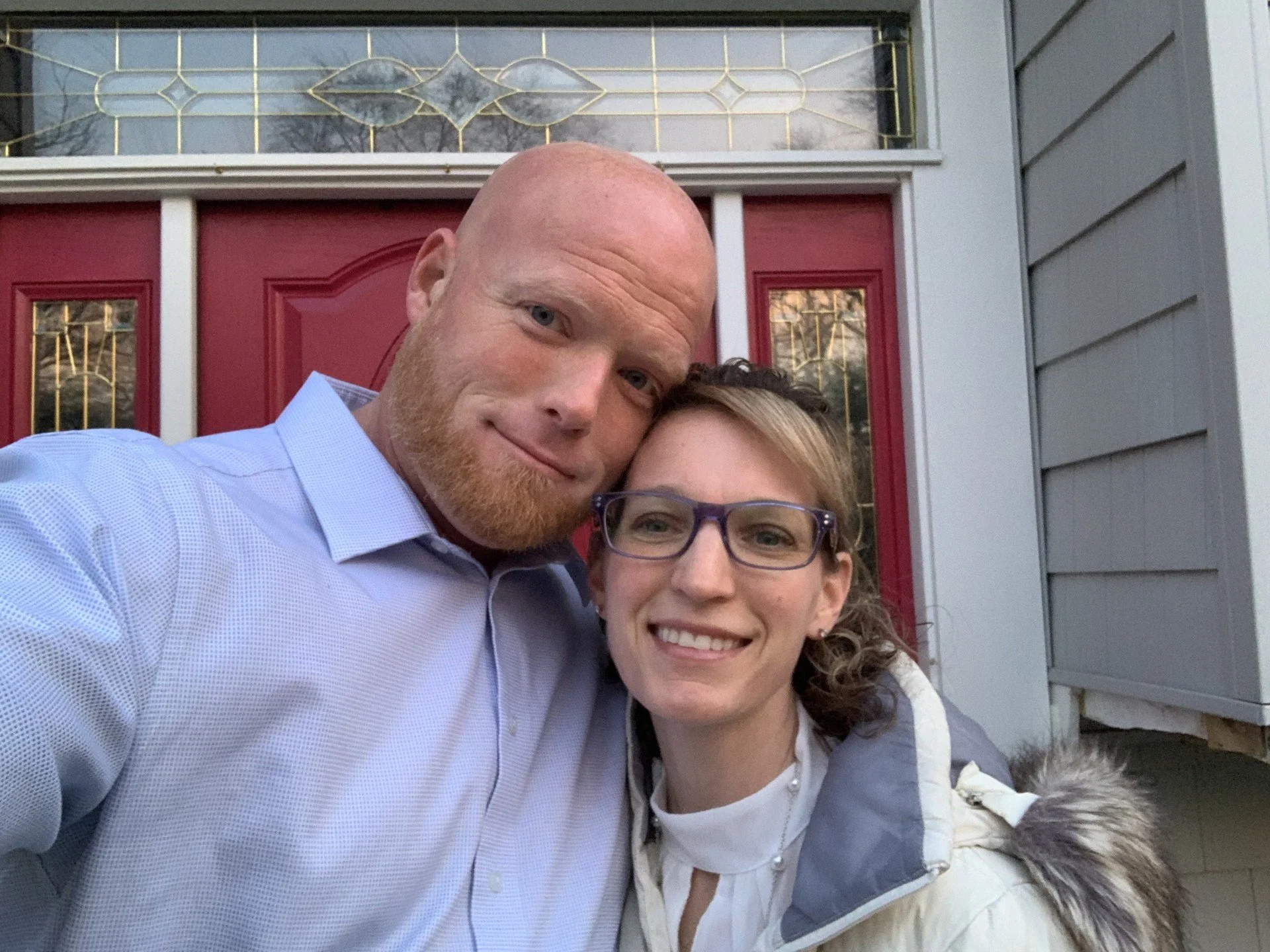 A man and woman taking a selfie outside a house with a red door and gray siding.