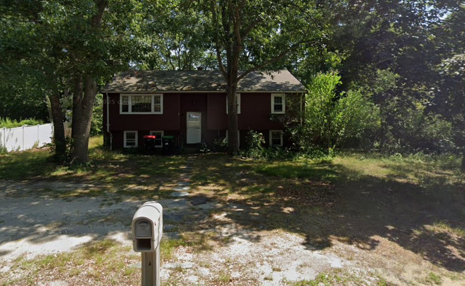 A small house with a dark red exterior, surrounded by trees, with a white fence to the left and a mailbox in the front yard.