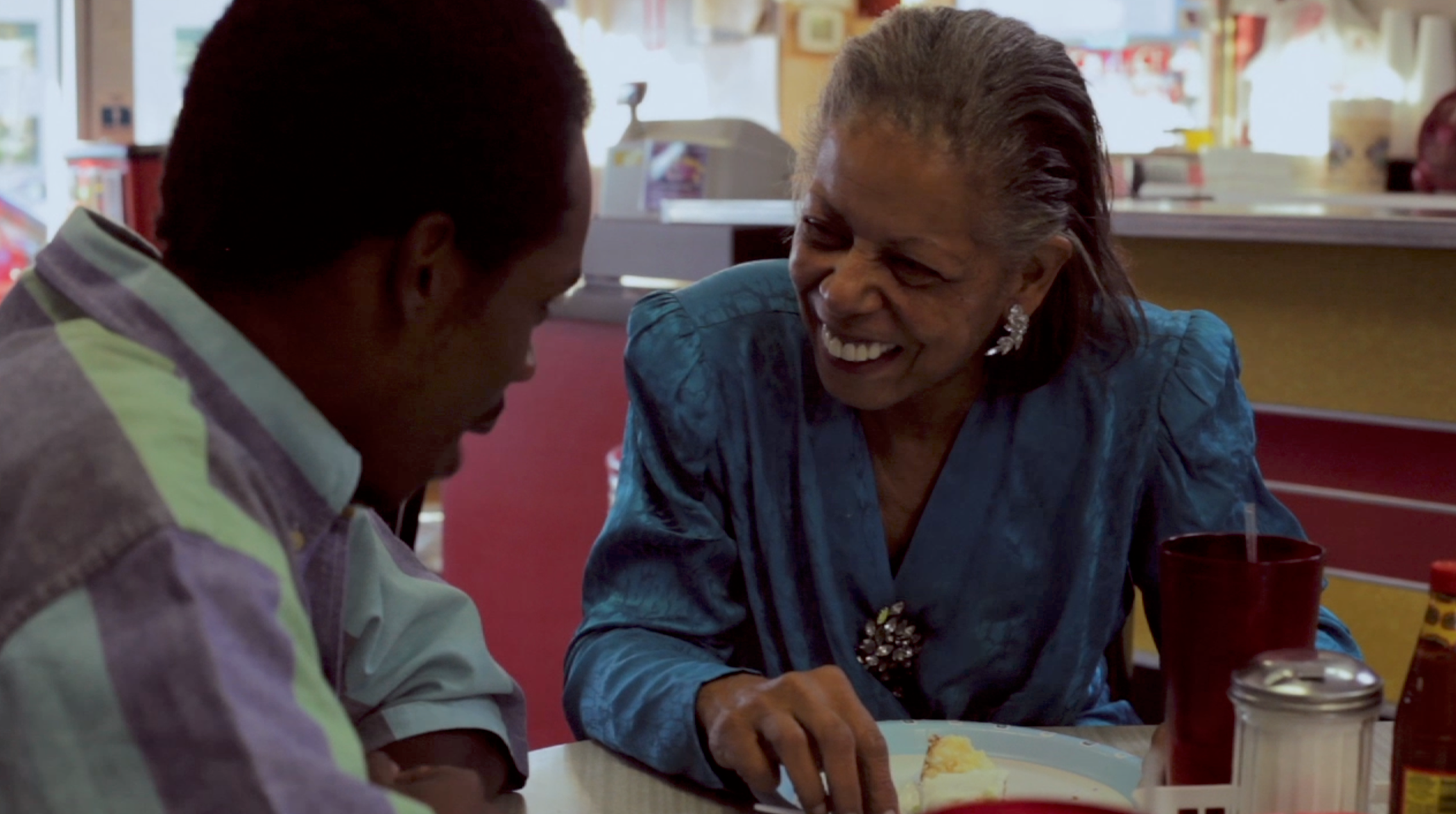 An elderly woman with gray hair and earrings laughing and smiling while sharing a meal with a man in a diner or restaurant.
