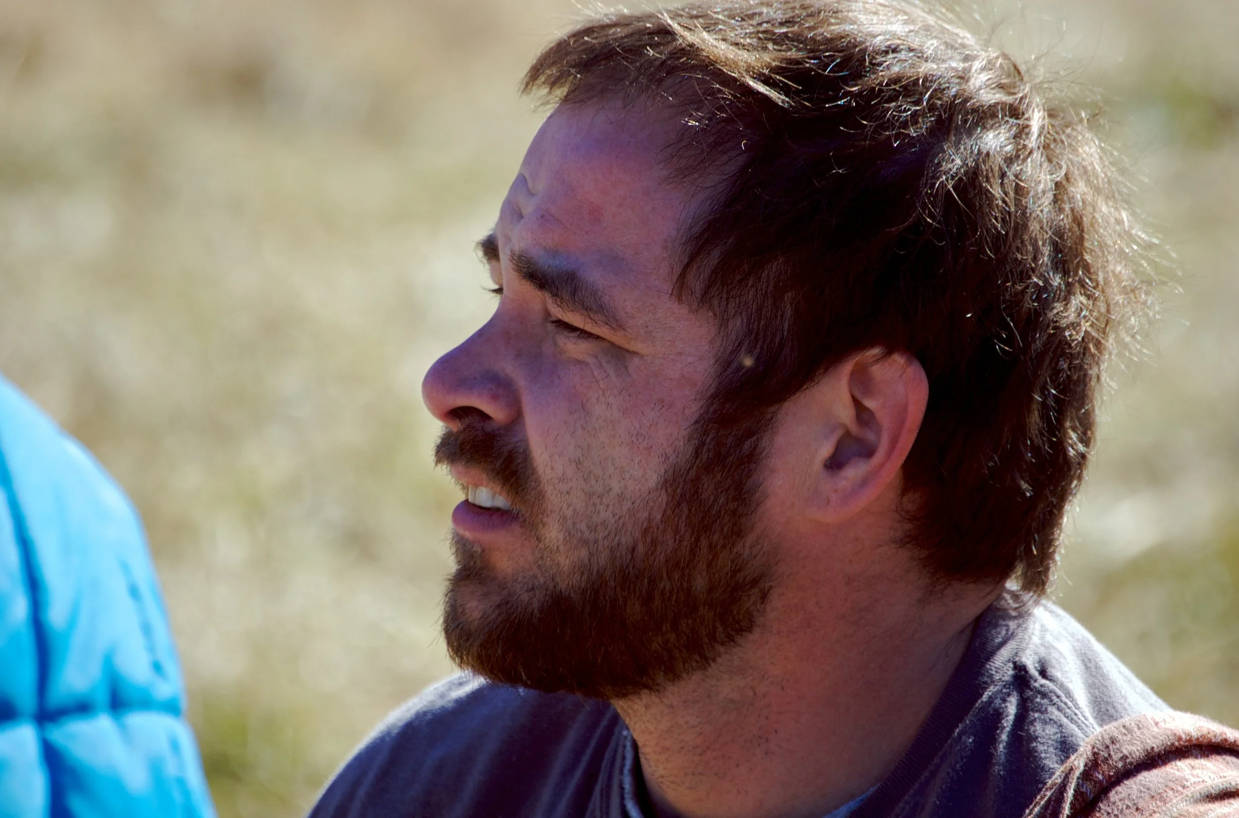 A man with a beard and short brown hair outdoors, squinting or grimacing, with a blurred natural background.