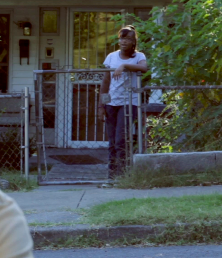 A girl standing on a porch behind a metal gate, wearing sunglasses and a white t-shirt, with greenery and a house in the background.