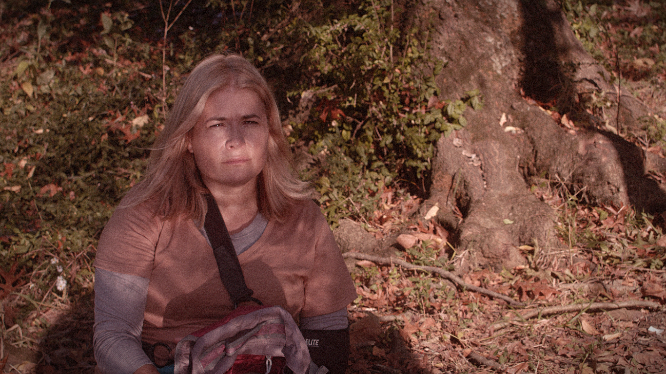 Young girl sitting outdoors on the ground among fallen leaves and branches, looking serious or concerned, with trees and foliage in the background.