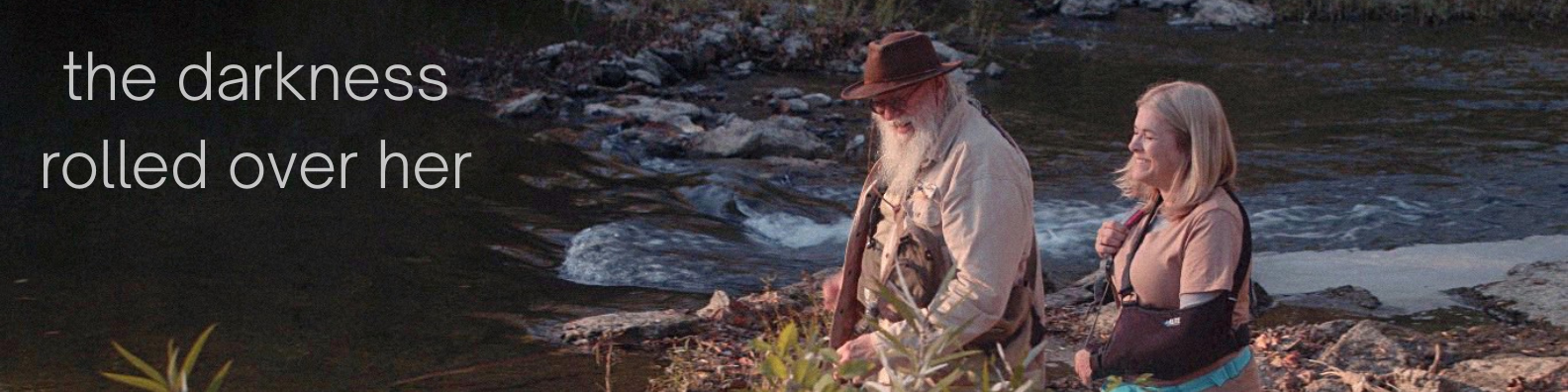 An elderly man with a beard and wearing a hat and sunglasses, and a woman with blonde hair, are standing by a riverbank outdoors. The man is looking down while the woman is smiling and looking at him.