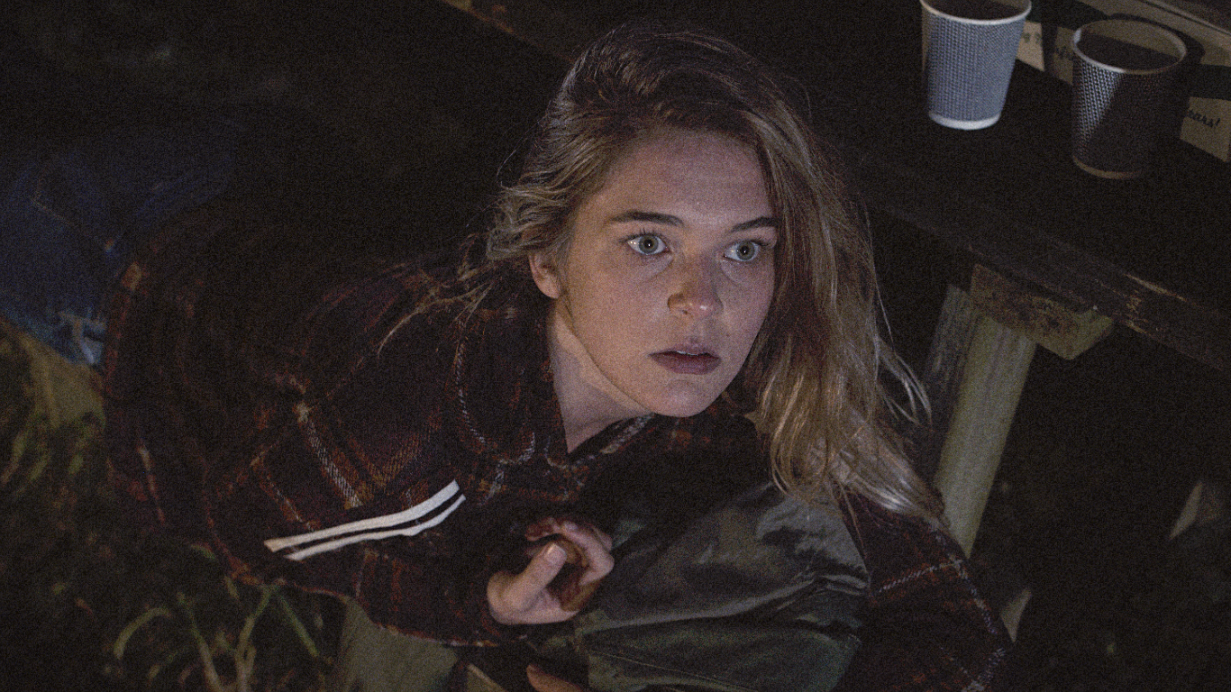 A young woman with wavy blonde hair and blue eyes looking up with a surprised or concerned expression in a dimly lit room with a table, disposable cups, and cups on the shelf behind her.