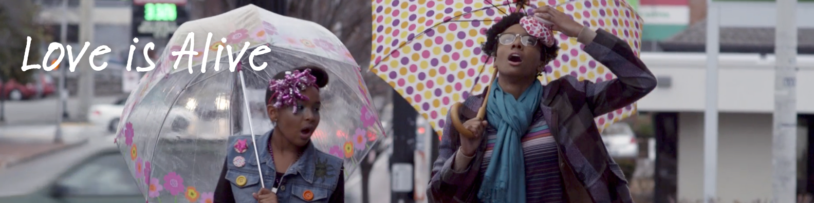 Two women walking with umbrellas on a rainy day, one with pink braids and bubblegum and the other with curly hair and glasses, with the text "Love is Alive" overlaid.