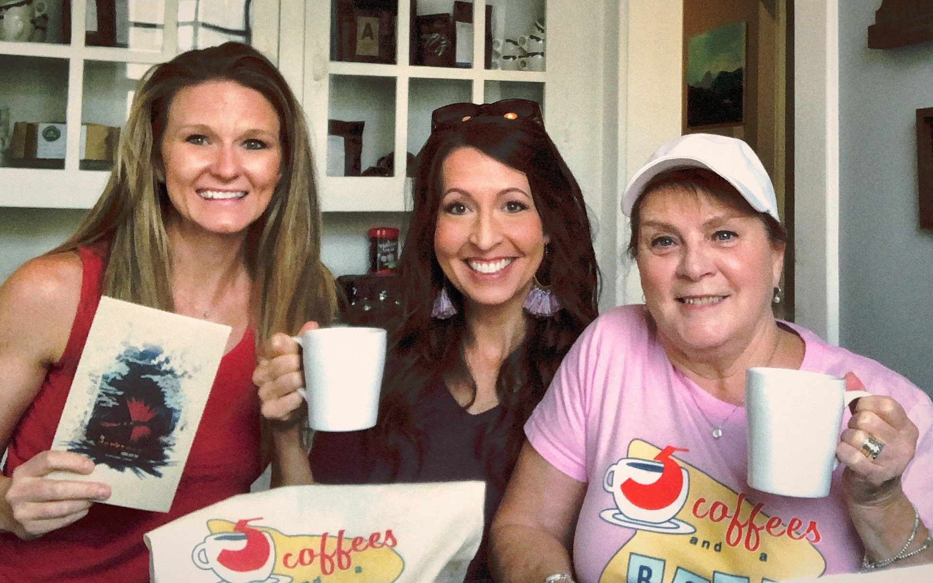 Three women smiling and holding white coffee mugs, sitting together in a cozy kitchen or cafe setting with white cabinets and various decorations in the background.