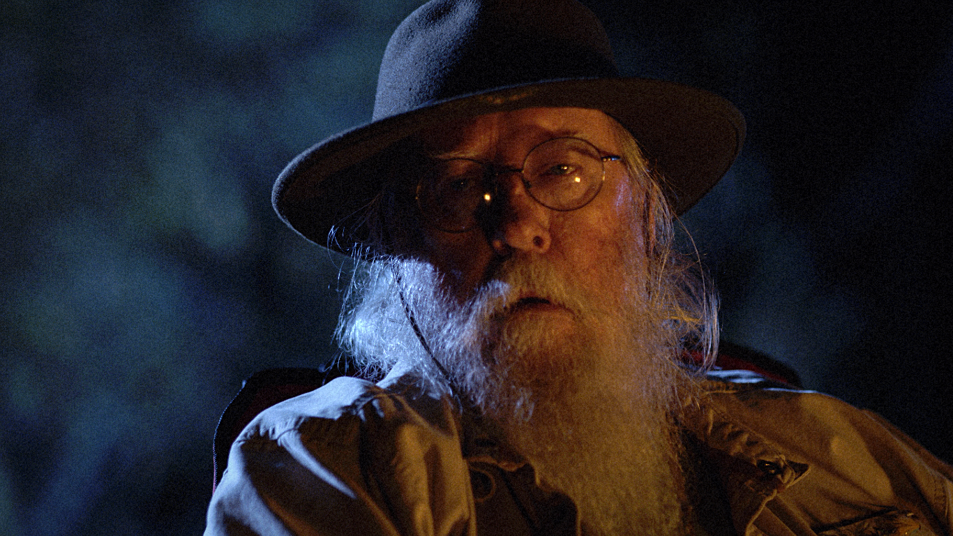 Close-up of an older man with a long white beard, glasses, wearing a wide-brimmed hat and a tan jacket, outdoors at night.
