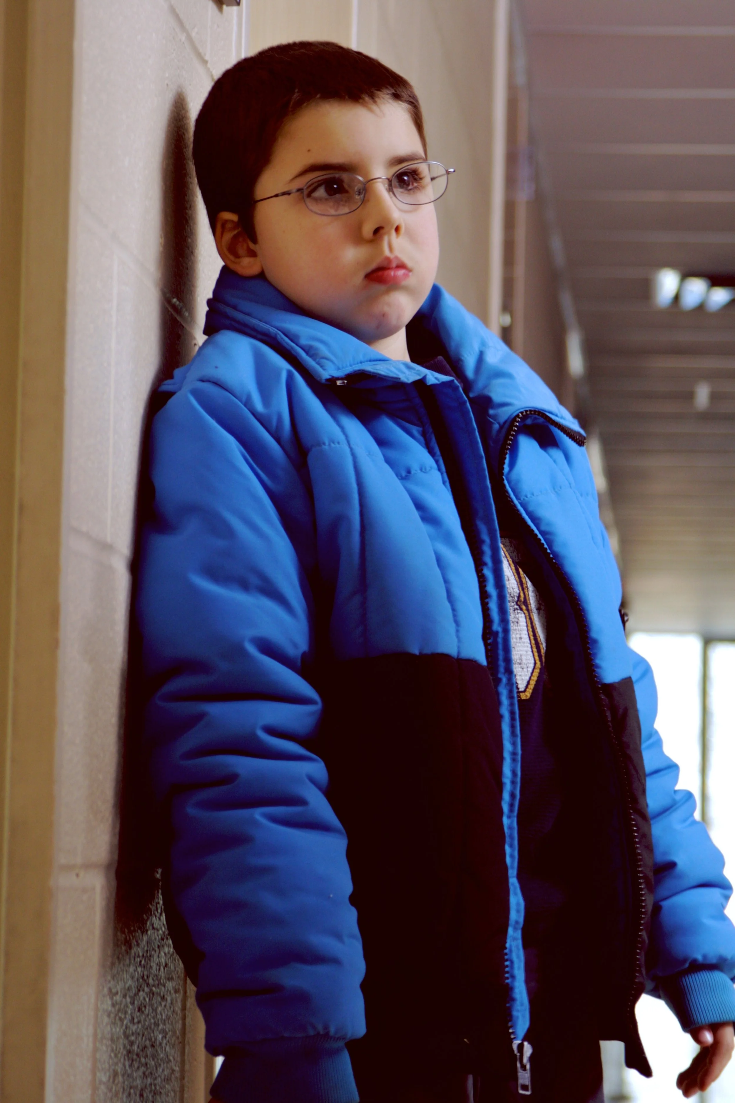 A boy with glasses wearing a blue puffer jacket leaning against a wall in a hallway, looking pensively to the side.