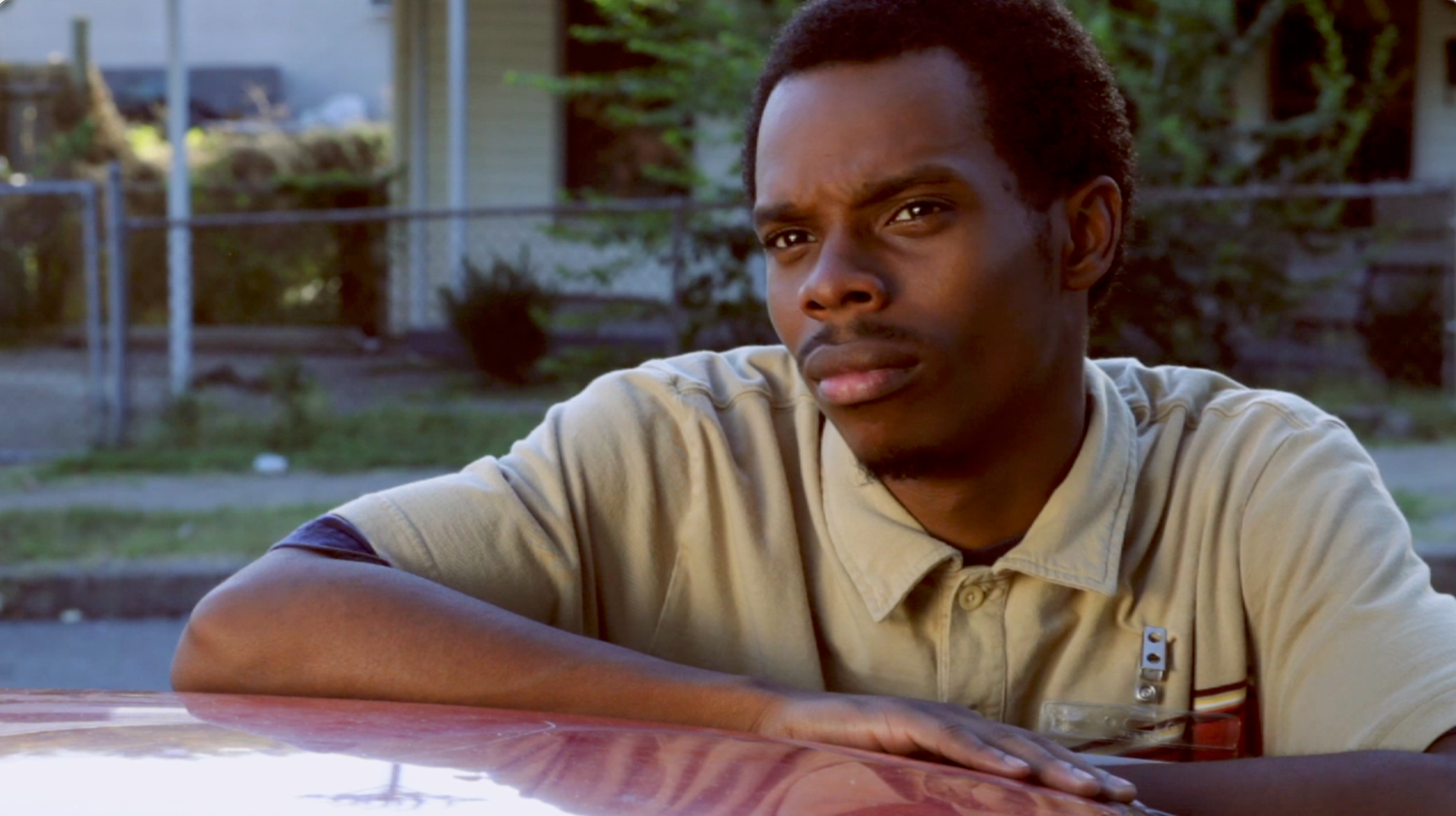A young man with short curly hair and dark skin, sitting outdoors at a table, looking slightly to the right with a serious expression. Background includes a chain-link fence, a grassy area, trees, and houses.