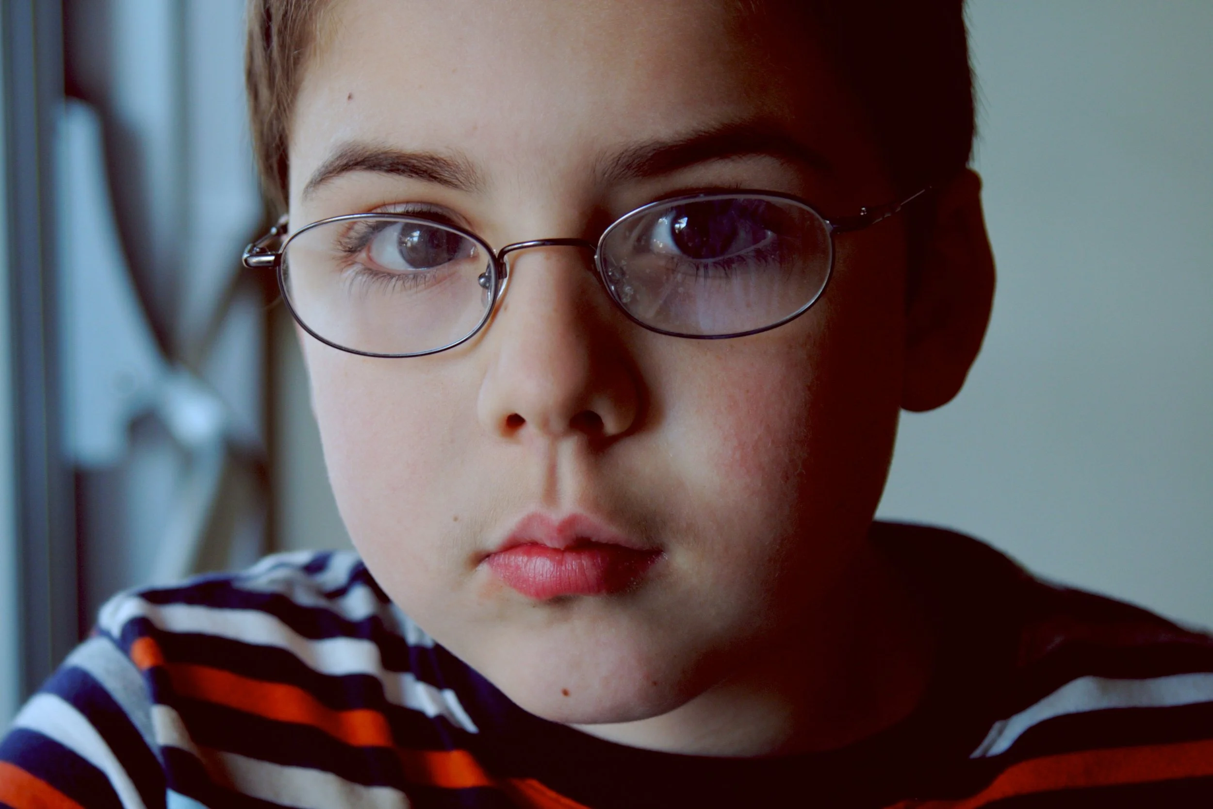 Close-up of a young person with glasses and short hair, wearing a striped shirt, looking into the camera.