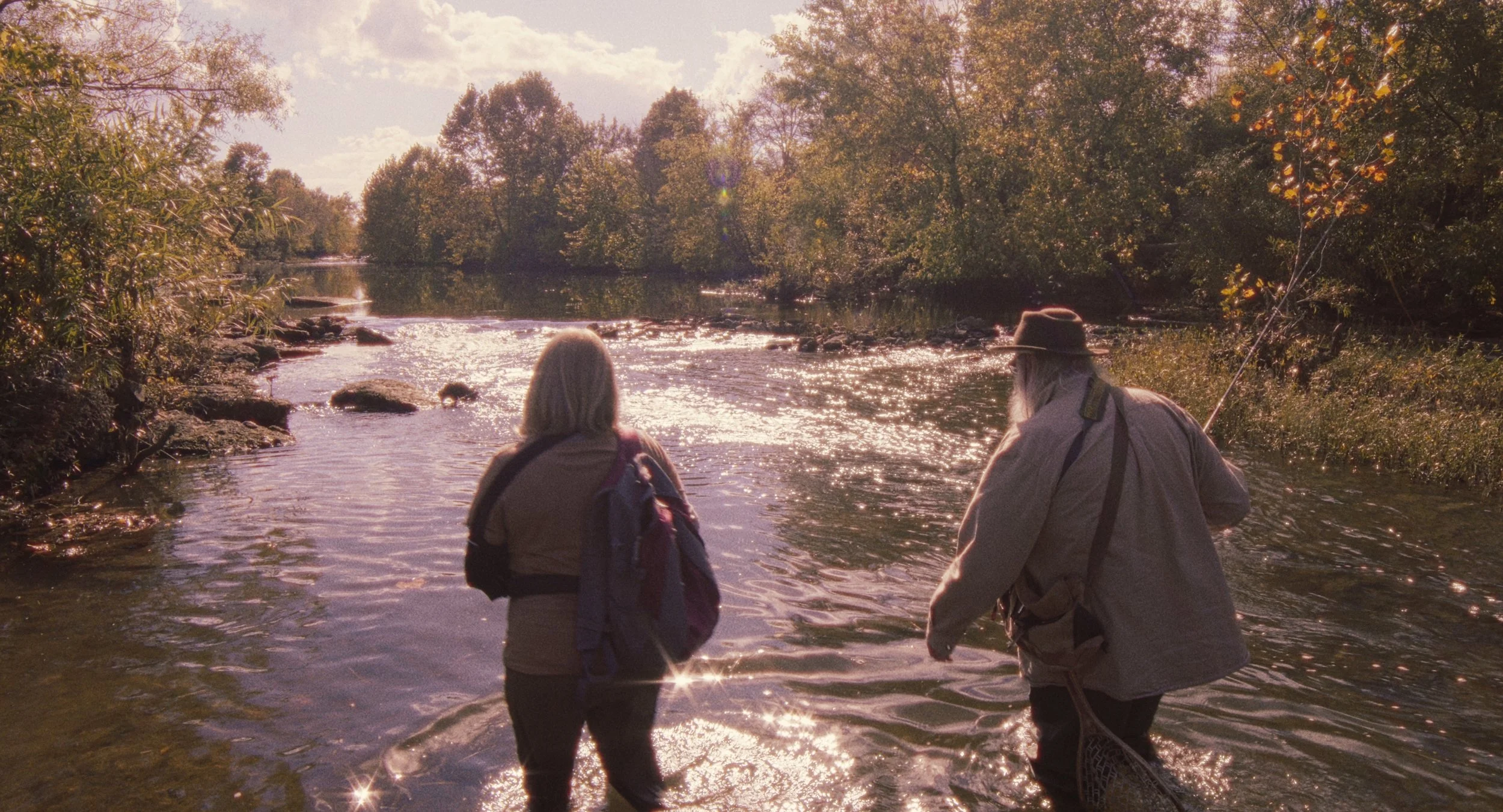 Two people, an older man with a fishing rod and hat, and a woman with a backpack, stand in a river surrounded by trees in warm sunlight.