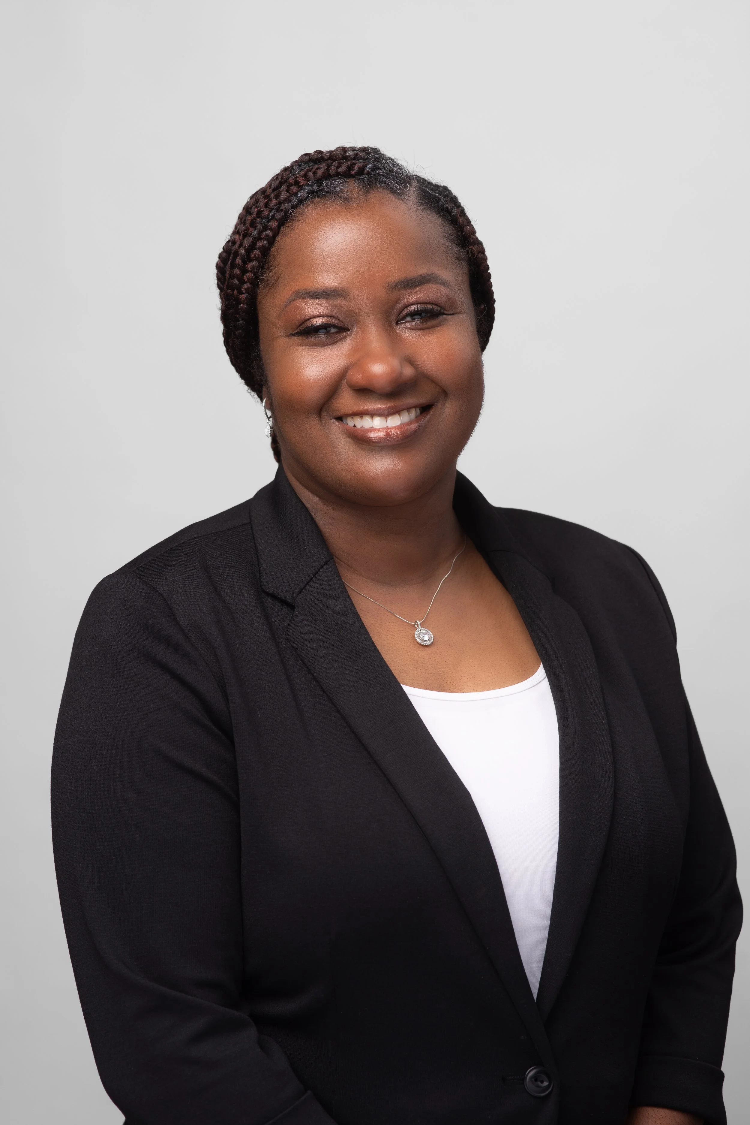 A professional woman with braids, wearing a black blazer and a white top, smiling against a plain light gray background.