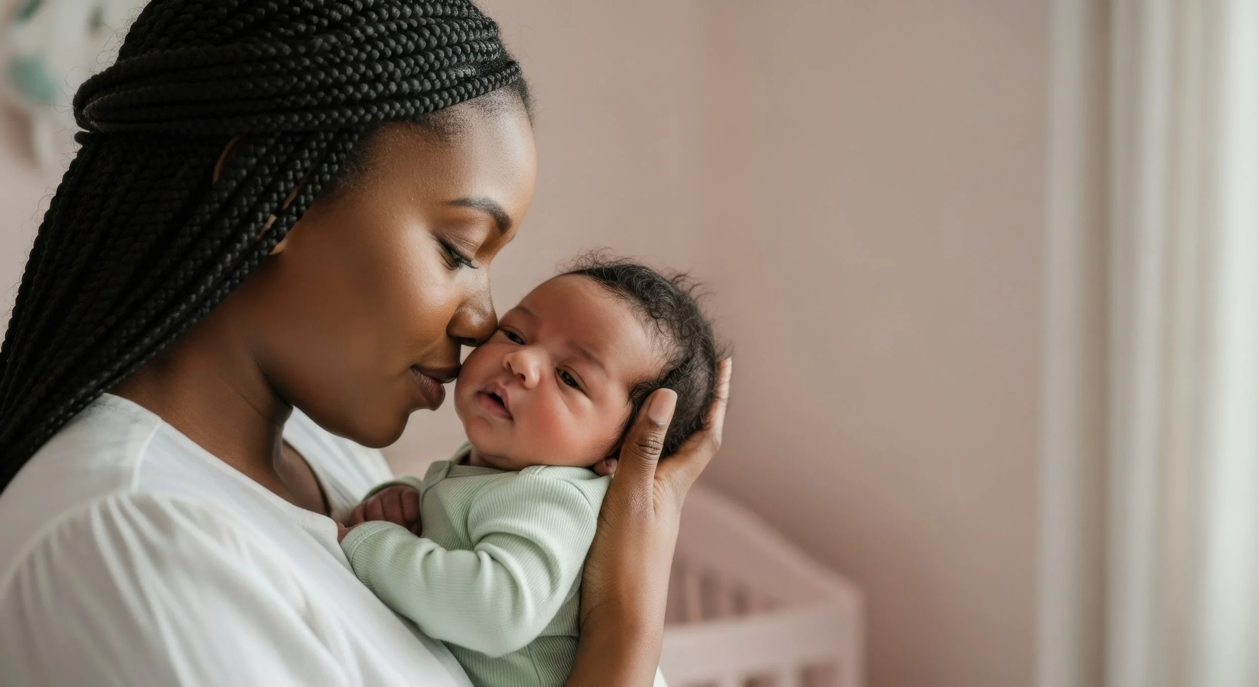 An African American woman with braided hair holding a baby close to her face, both with eyes closed, sharing a tender moment indoors.