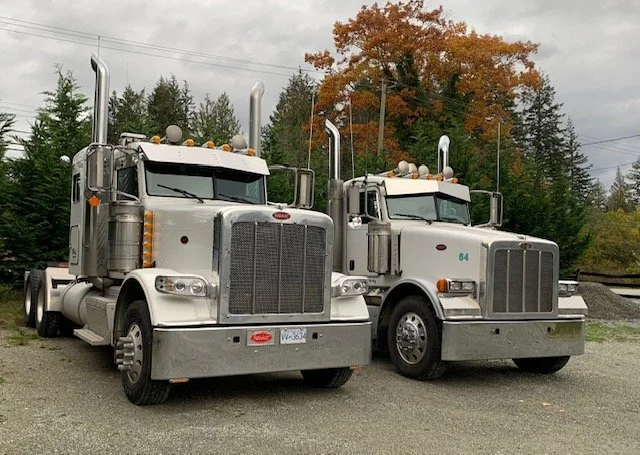 Two silver semi-trucks parked on a gravel surface with trees and autumn foliage in the background.