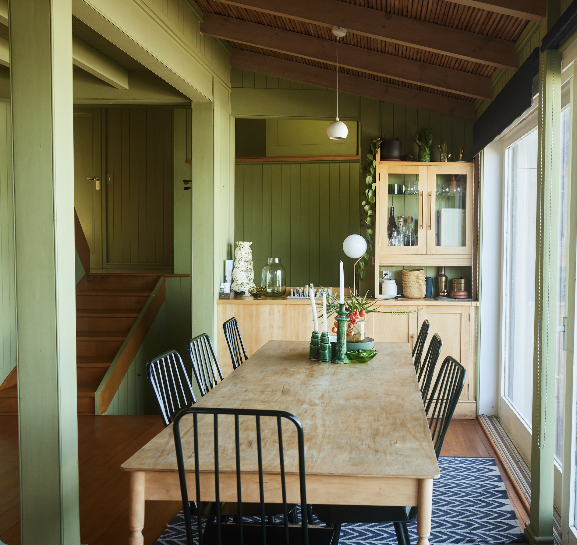 A wooden dining table with six black metal chairs, green cabinets, and a green wall with decorative vases, cactuses, and plants inside a bright room with glass doors.
