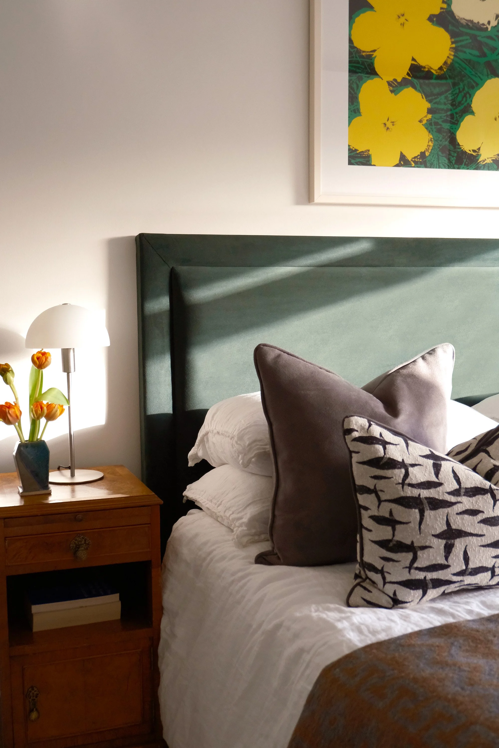 A bedroom with a white bed, a green headboard, pillows, a wooden bedside table with a lamp and flowers, and a framed yellow floral artwork on the wall.
