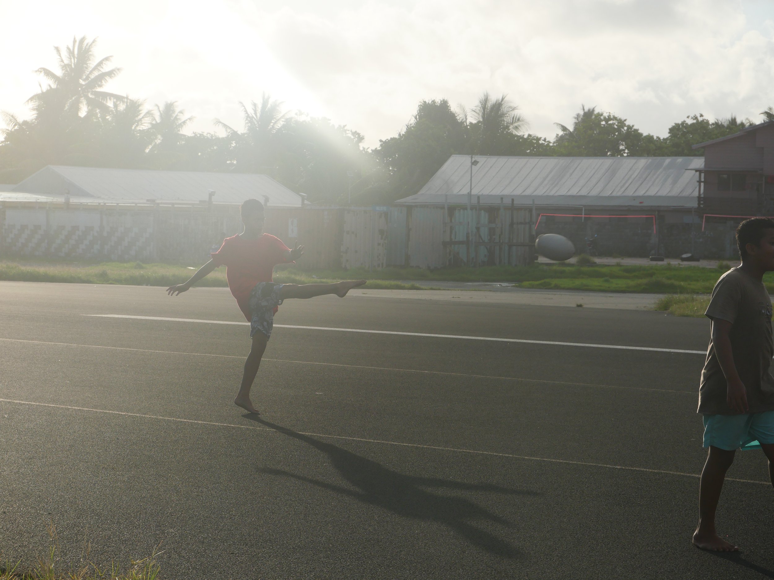 A young boy in a red shirt and patterned shorts performing a high kick on a paved street during the daytime, with two other children nearby and houses with metal roofs and palm trees in the background.