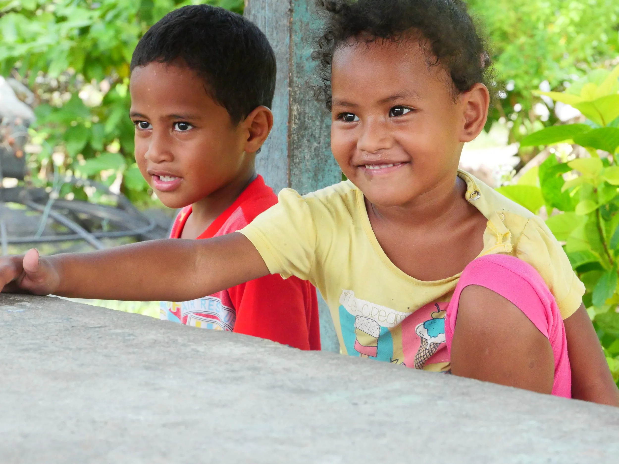Two smiling children, a girl in yellow and pink and a boy in red, playing outdoors behind a concrete barrier with green foliage in the background.