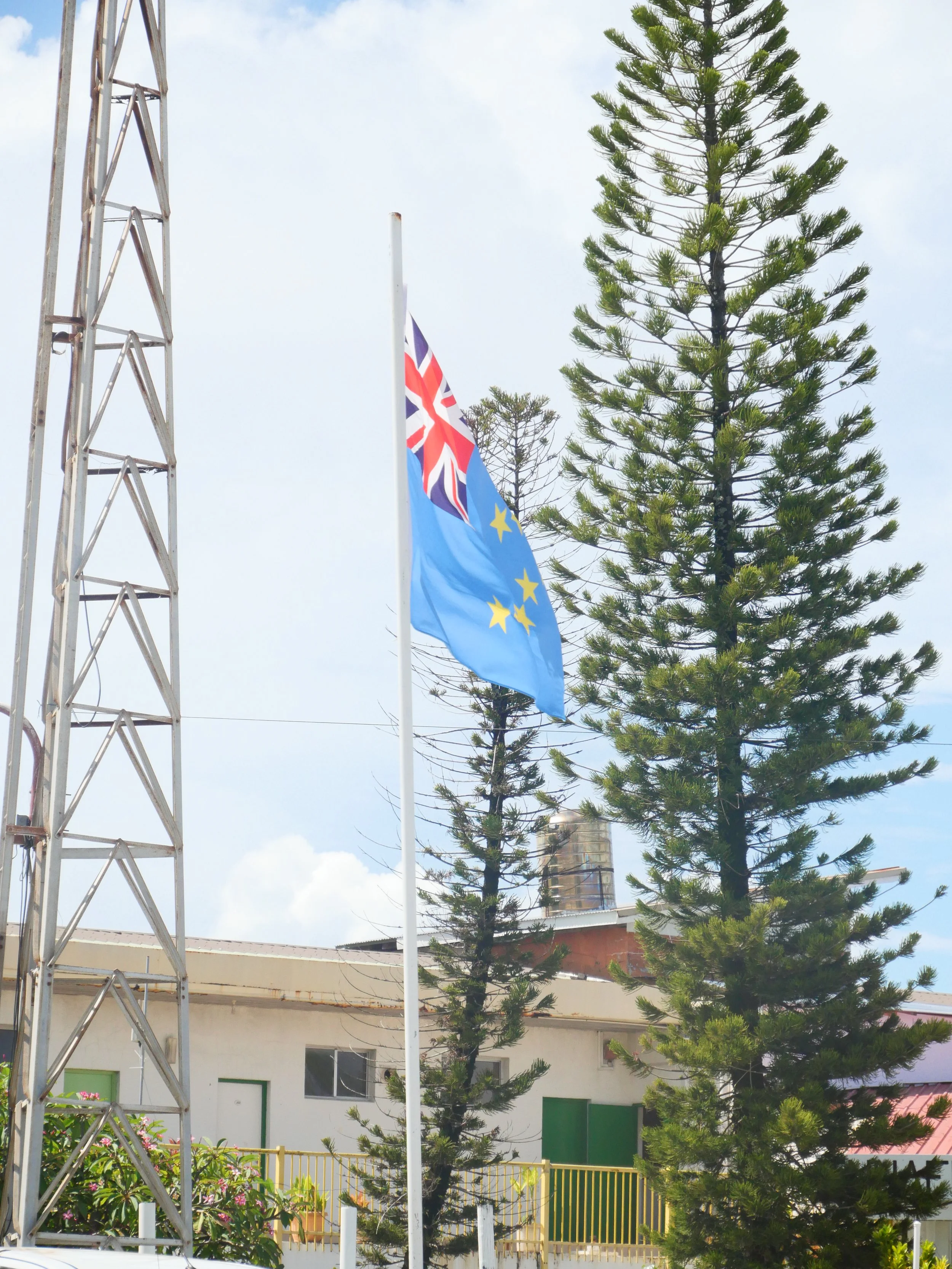 Flagpole with the New Zealand flag and a European Union flag in an outdoor area with trees and buildings in the background.