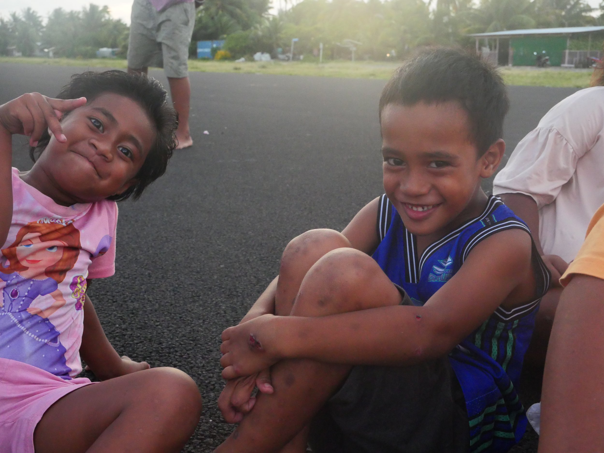Two children sitting on the ground outdoors, smiling at the camera. The girl on the left is wearing a pink shirt with a princess graphic and shorts, making a peace sign with her fingers. The boy on the right, wearing a blue sports jersey, has visible