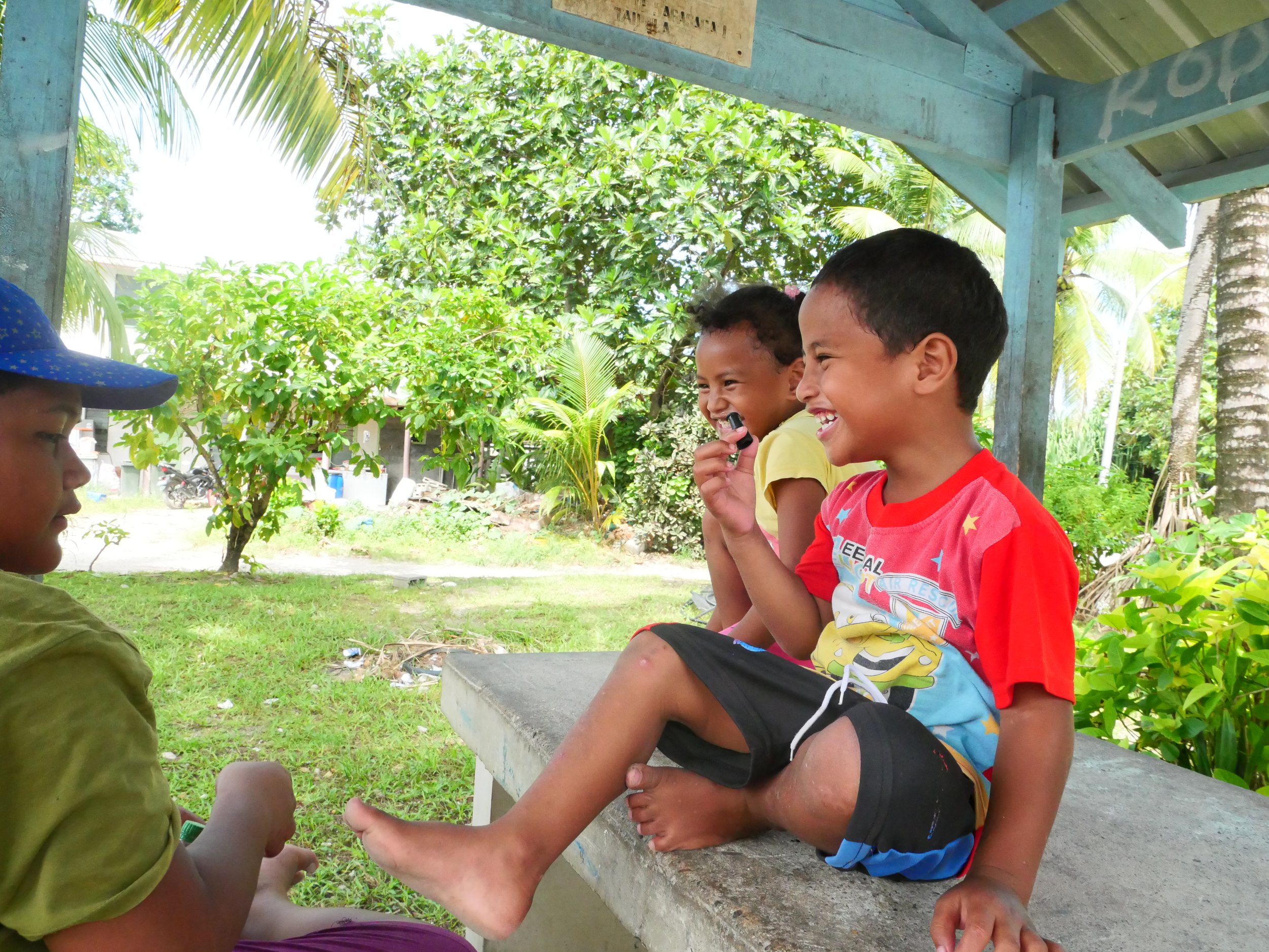Three children sitting on a concrete bench outdoors, laughing and playing together in a tropical area with lush greenery.