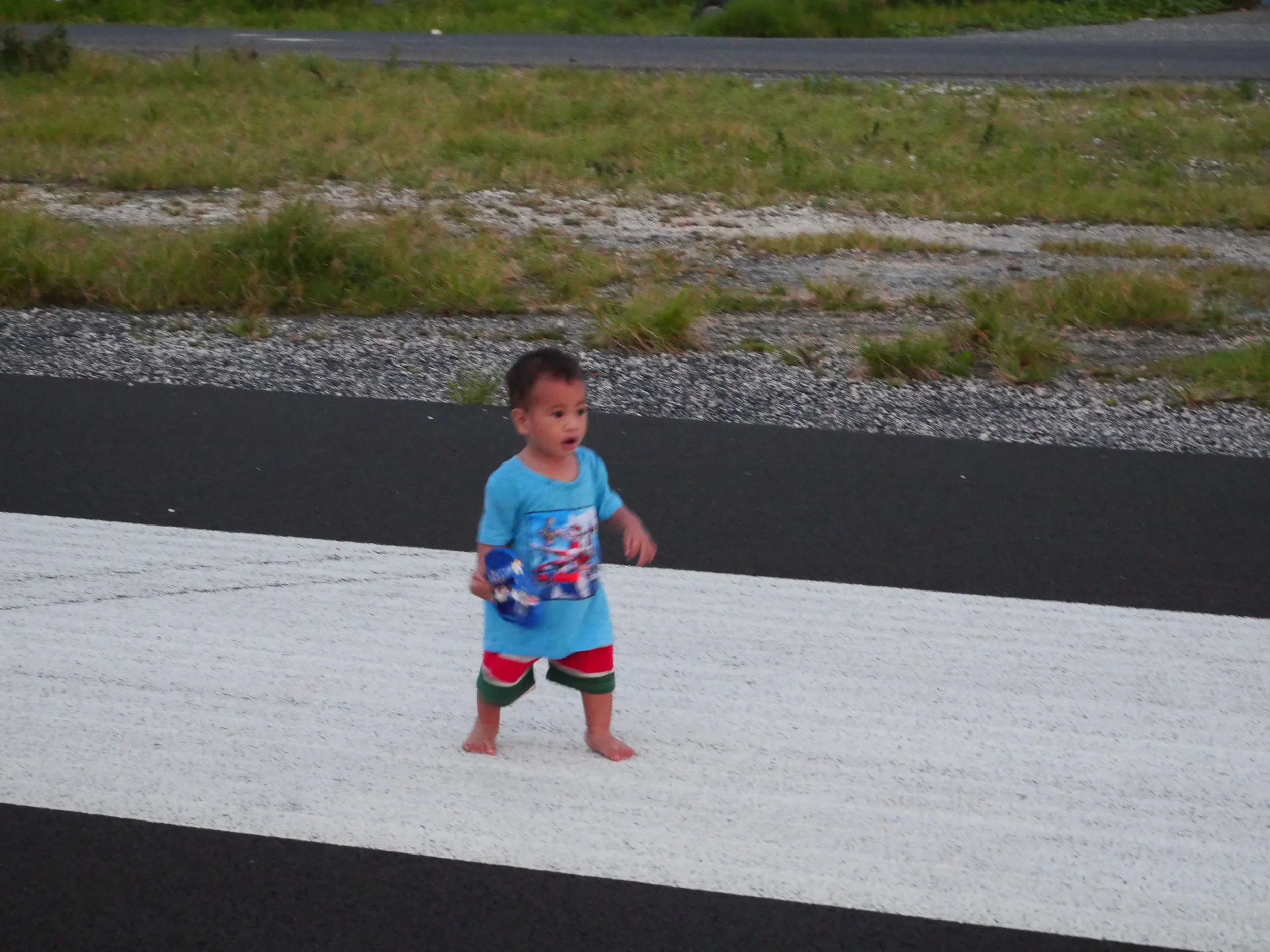 A young boy standing on a pedestrian crosswalk on the side of the road, wearing a blue shirt with a graphic and colorful shorts, with a surprised or curious expression.