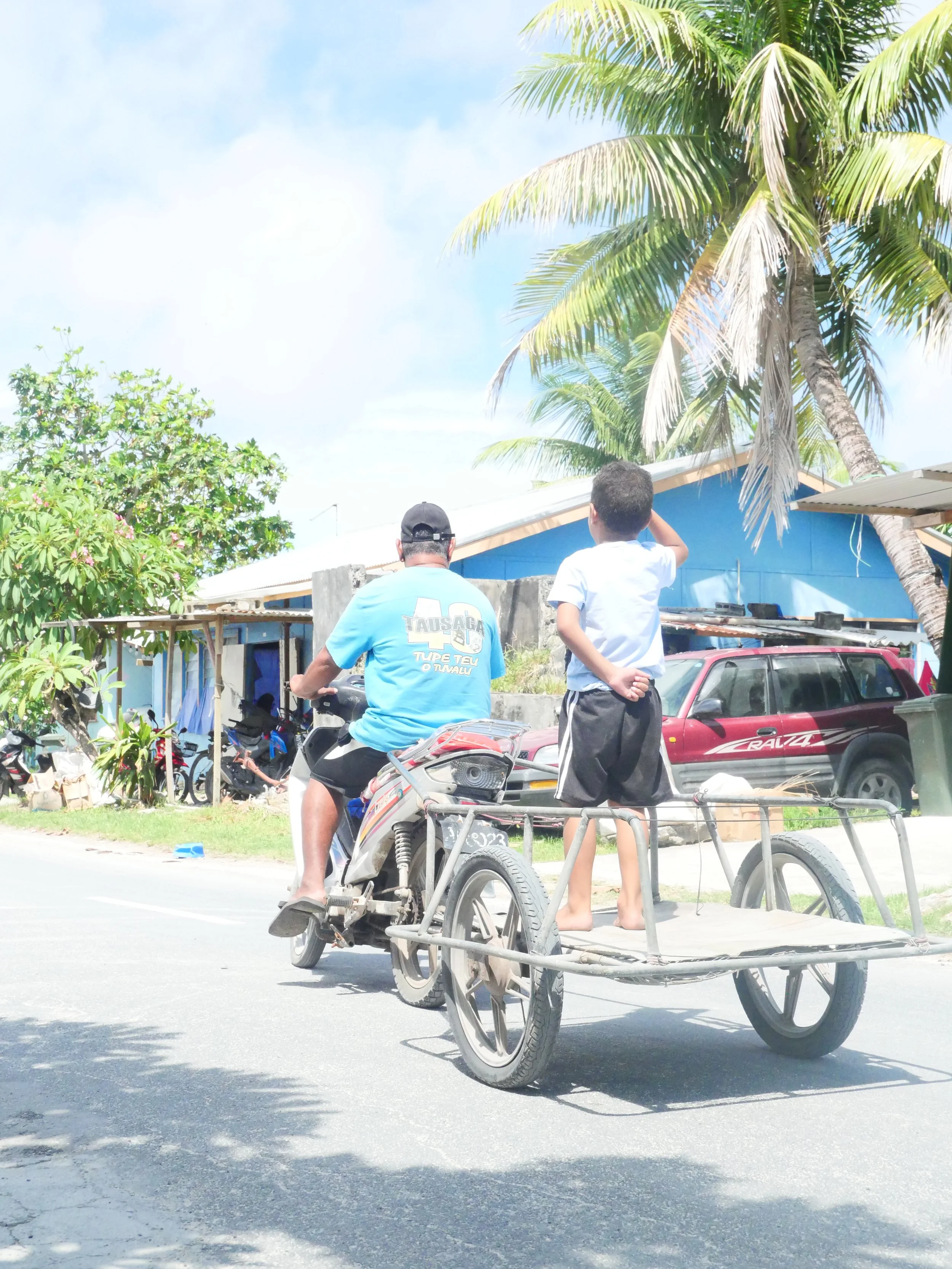 A man riding a motorcycle with a cart attached, carrying a boy who is standing on the cart and looking down the road, with palm trees, a blue house, and parked cars in the background on a sunny day.