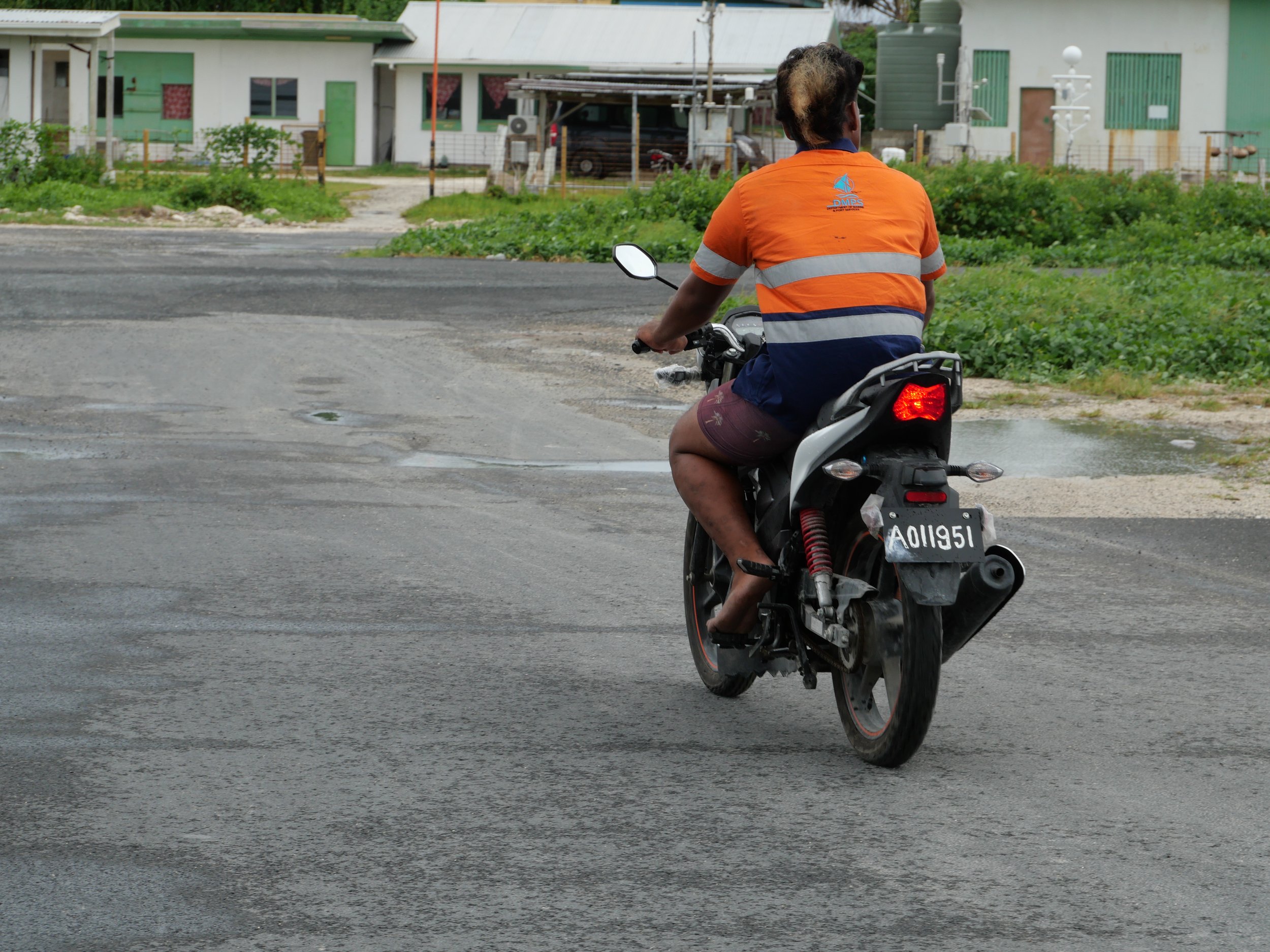 A man wearing an orange safety vest riding a motorcycle on a paved road in a rural area.
