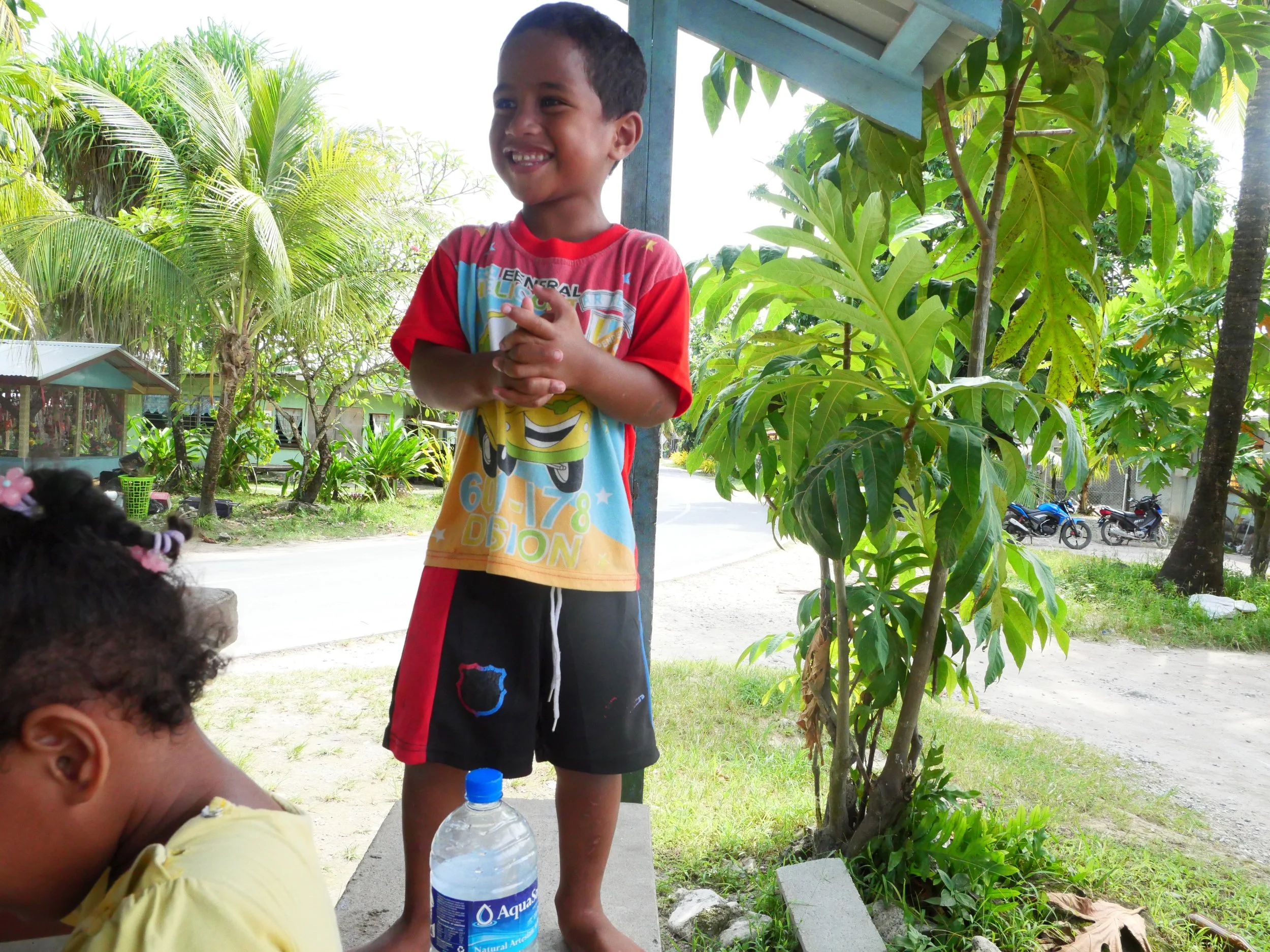 A smiling young boy standing outdoors on a sunny day, wearing a colorful cartoon t-shirt and black shorts with red sides. He is clasping his hands and looks happy. There is a girl in yellow with curly hair near the bottom left corner and a plastic wa