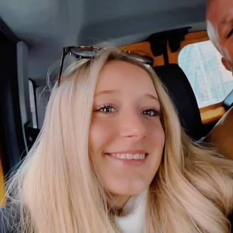 A young woman with long blonde hair smiling inside a vehicle.