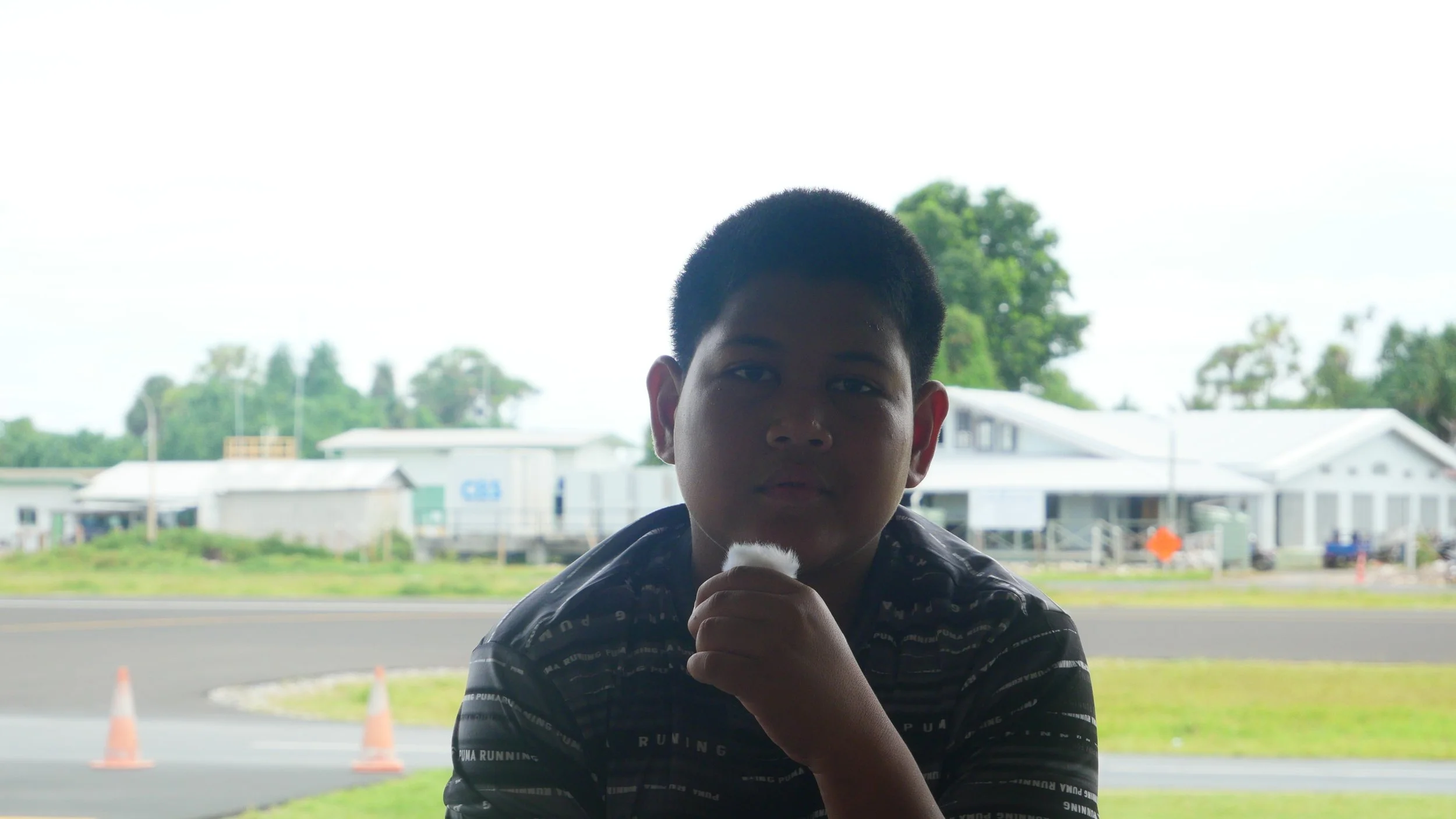 A young boy with short black hair and wearing a dark patterned shirt, sitting outdoors near a road, with a background of white buildings, trees, and orange traffic cones.