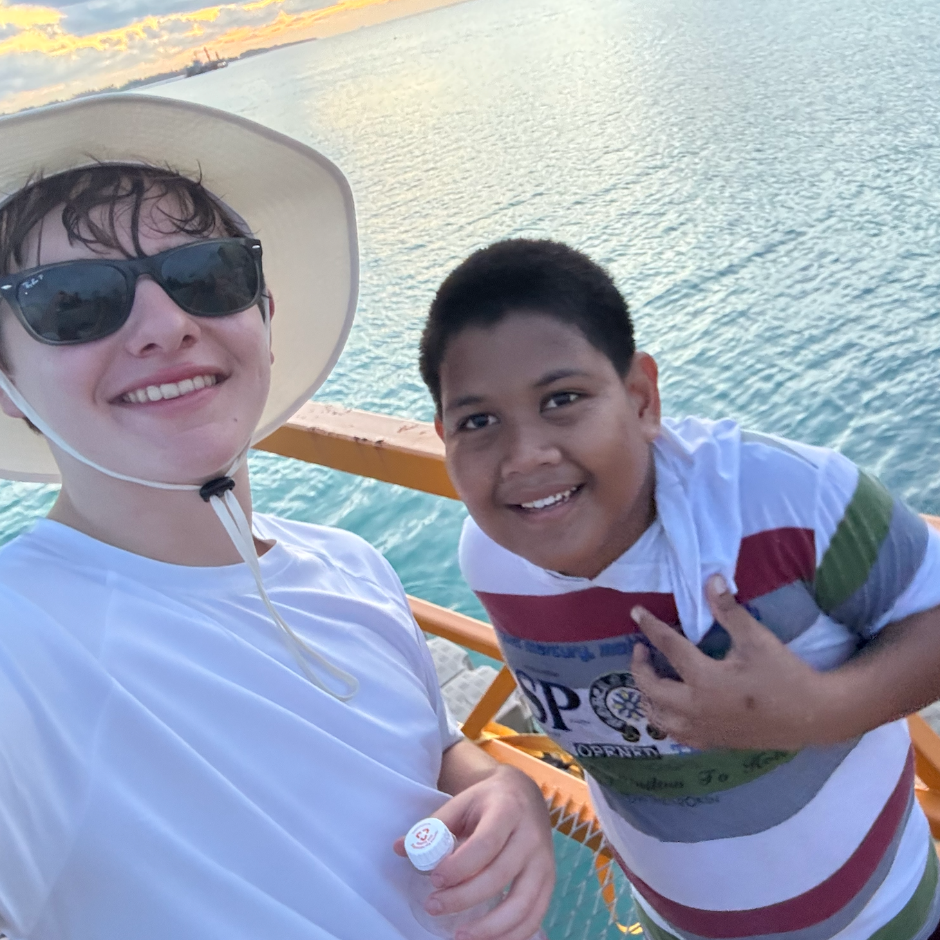 Two smiling people, one wearing sunglasses and a hat, the other with short hair, on a boat near water during sunset.