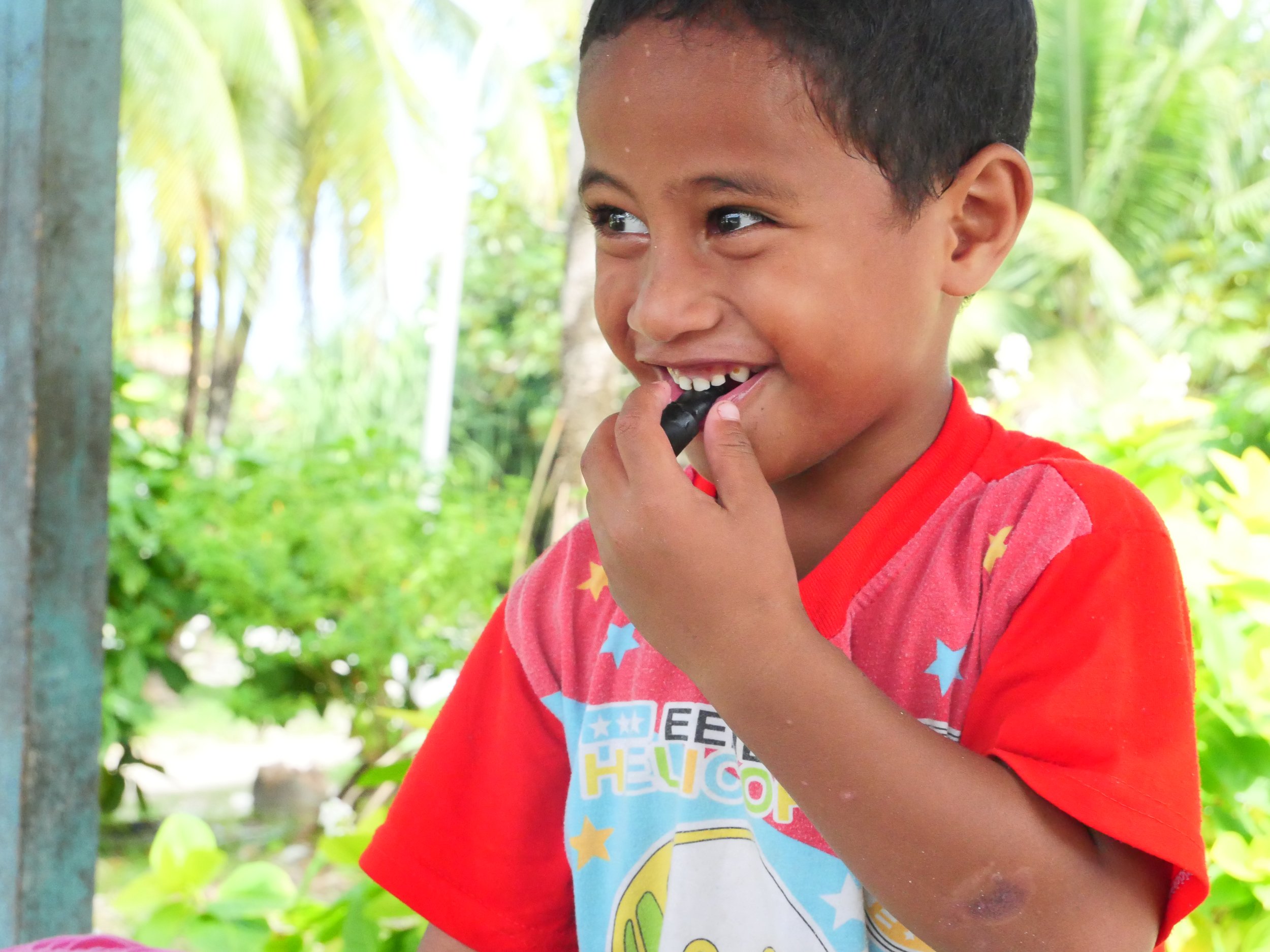 A young boy in a red shirt with colorful stars, smiling and biting his finger outdoors with green foliage and trees in the background.