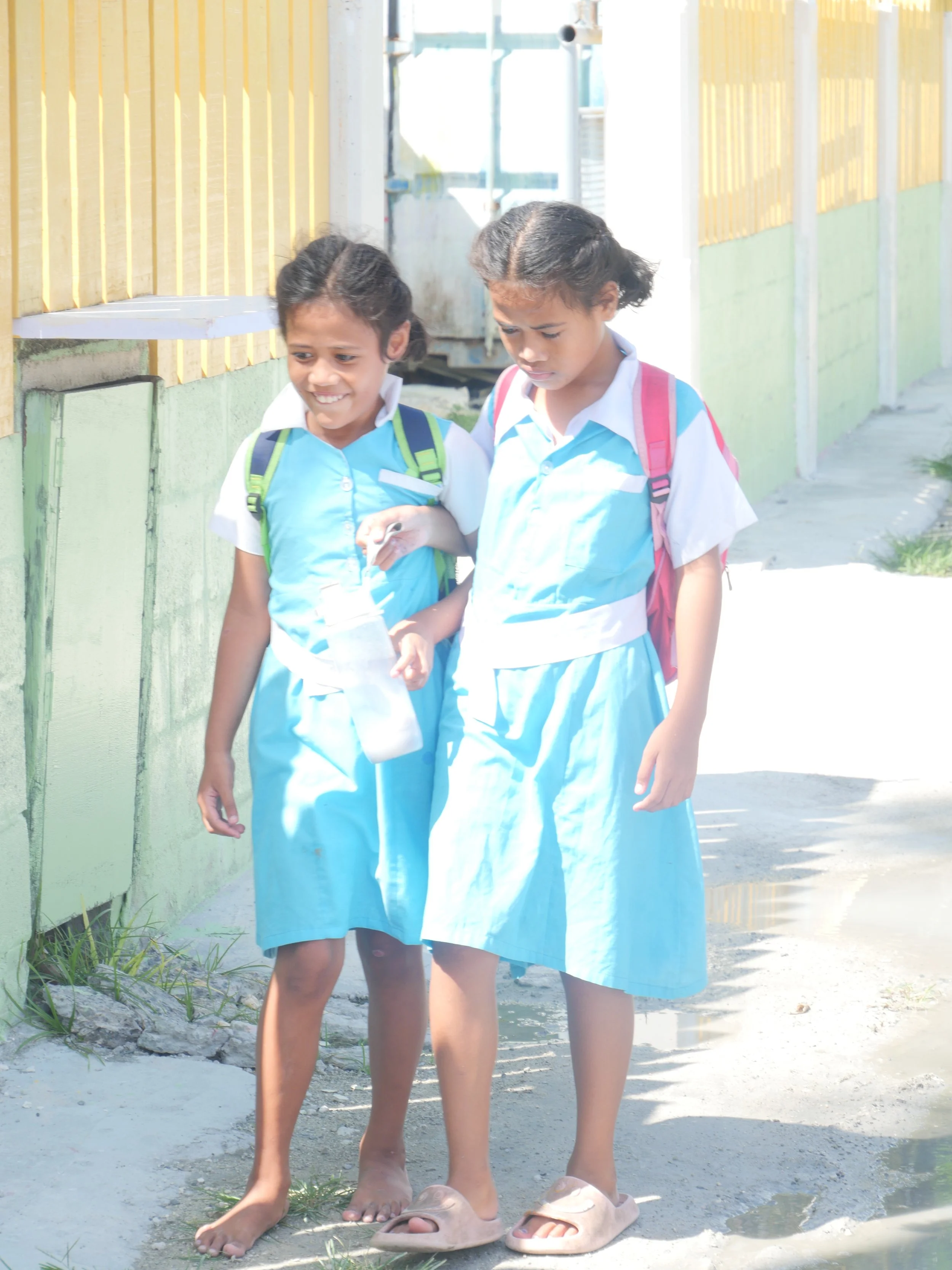 Two young girls in school uniforms walking outdoors holding hands and smiling.