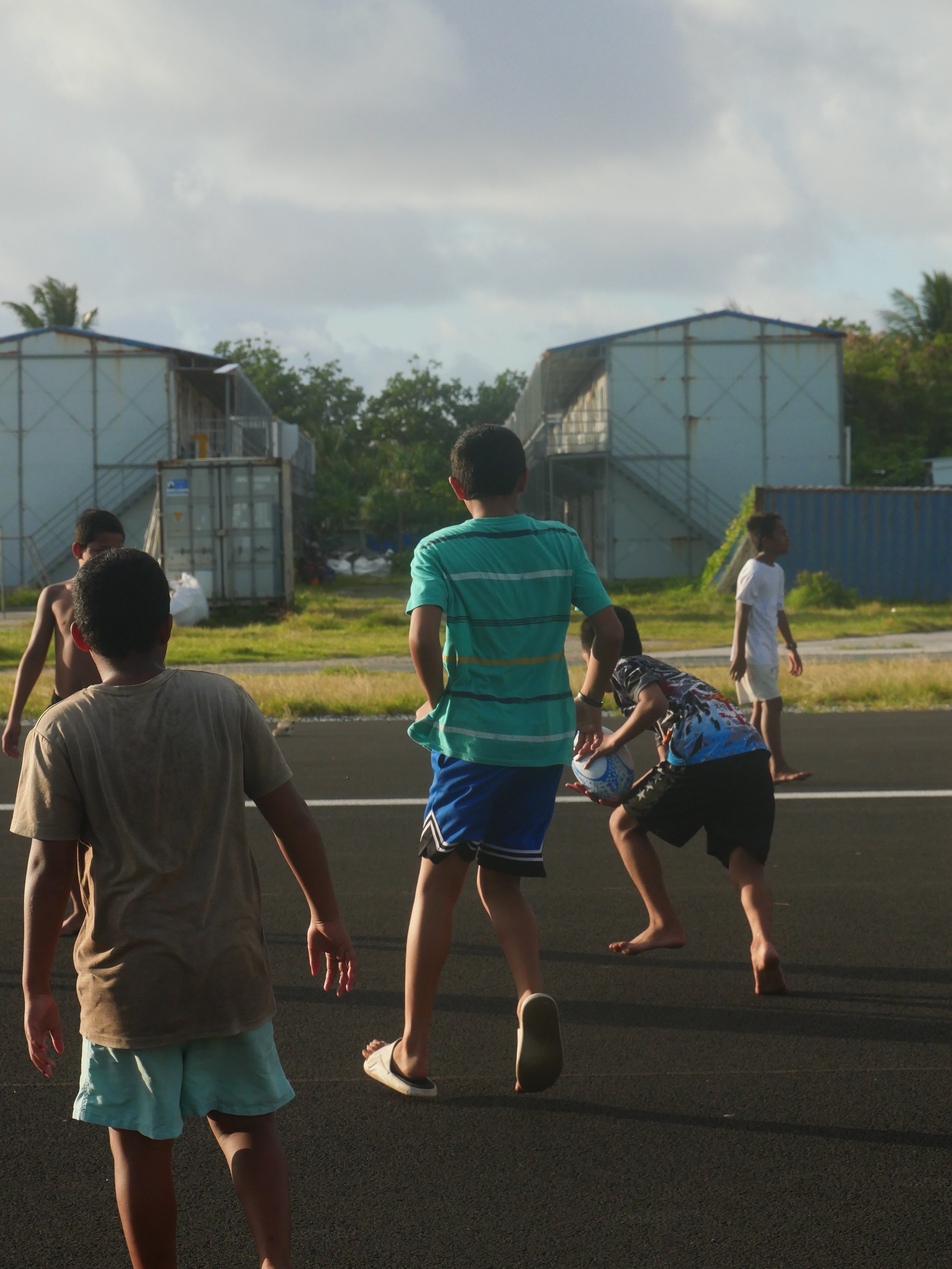 Children playing football outdoors on a blacktop surface in a tropical setting with buildings and trees in the background.