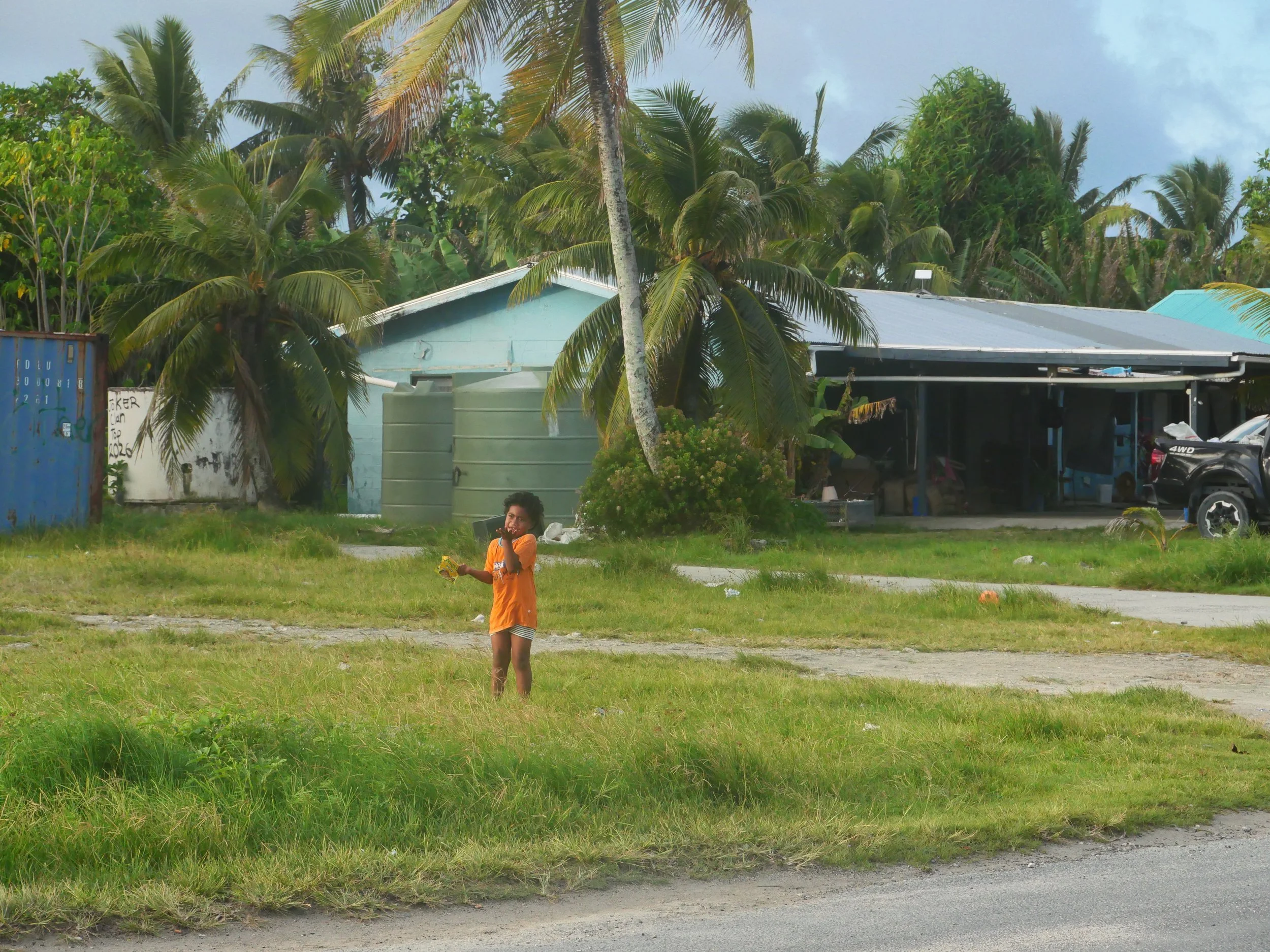 A young girl in an orange shirt and striped shorts standing on grass while talking on a cellphone and holding a yellow toy. Behind her are palm trees and a house with a grey metal roof, a car parked nearby, and a blue shipping container.