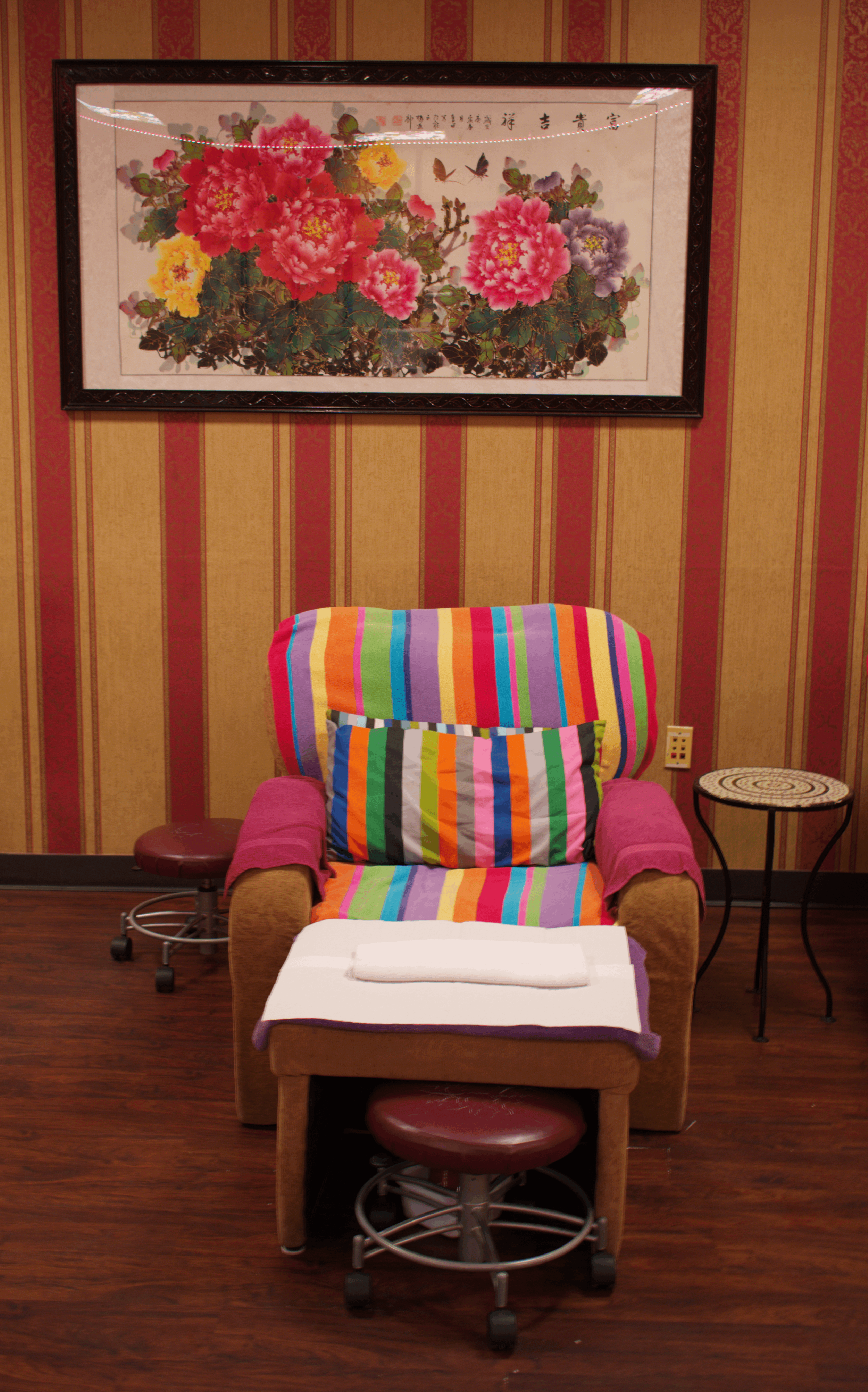 A massage or pedicure chair draped with colorful striped fabric and a towel, located in a room with wood flooring, decorated with a framed floral painting, a small round table, and a rolling stool.