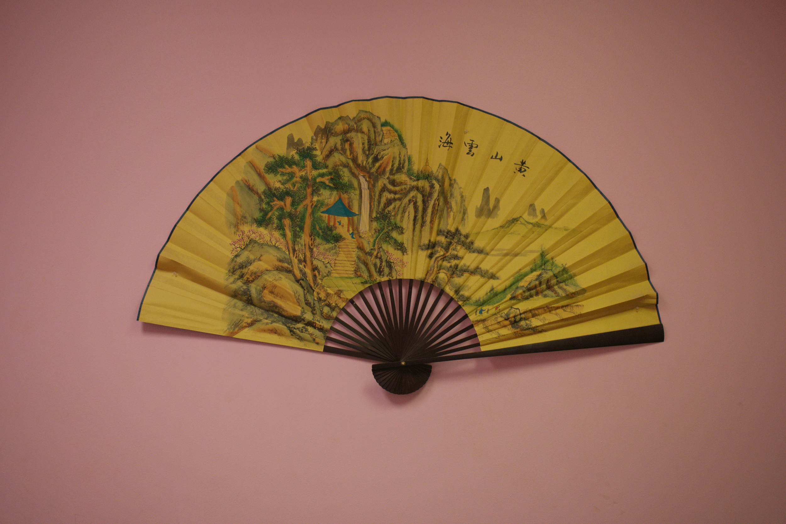 Traditional Asian folding fan with a painted landscape of mountains, waterfalls, trees, and a small building, set against a pink background.
