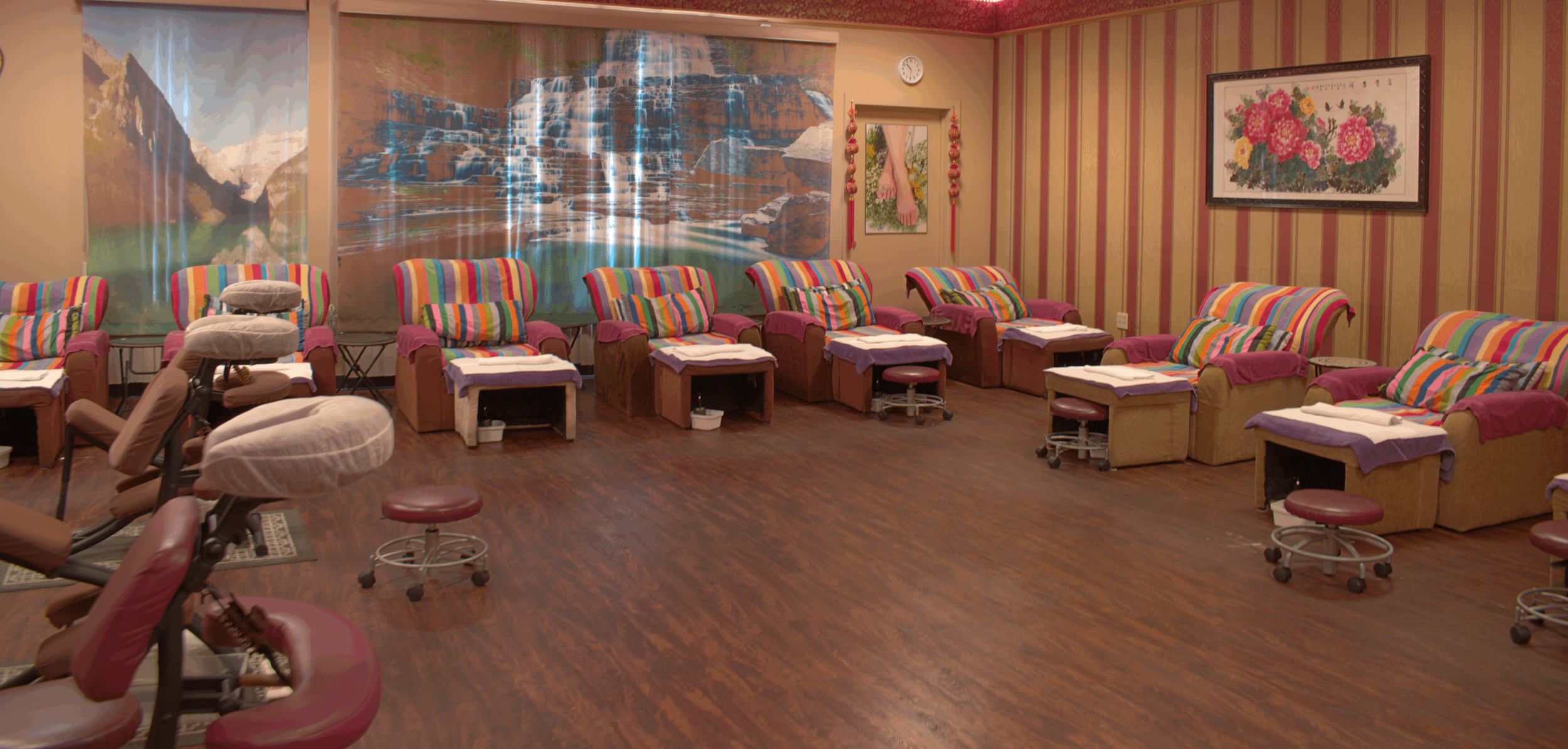 Spa or relaxation room with multiple massage chairs covered with colorful striped towels, small tables beside each chair, a large scenic waterfall mural on the wall, floral artwork, and a clock on the wall.